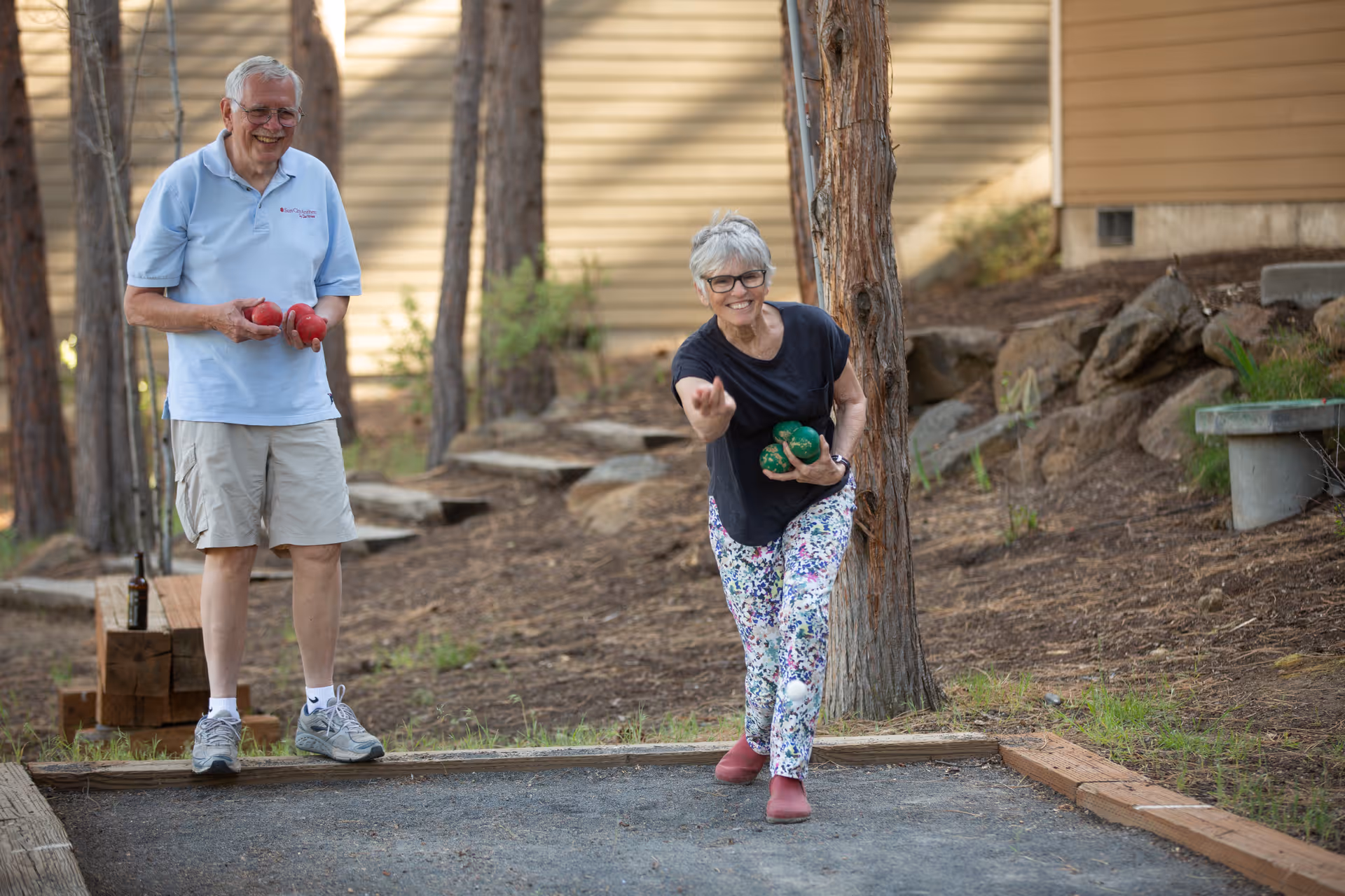 An elderly woman is playing bocce ball outdoors, throwing a ball while holding two others. An elderly man stands nearby smiling, holding two bocce balls. They are in a wooded area with trees and a building in the background.
