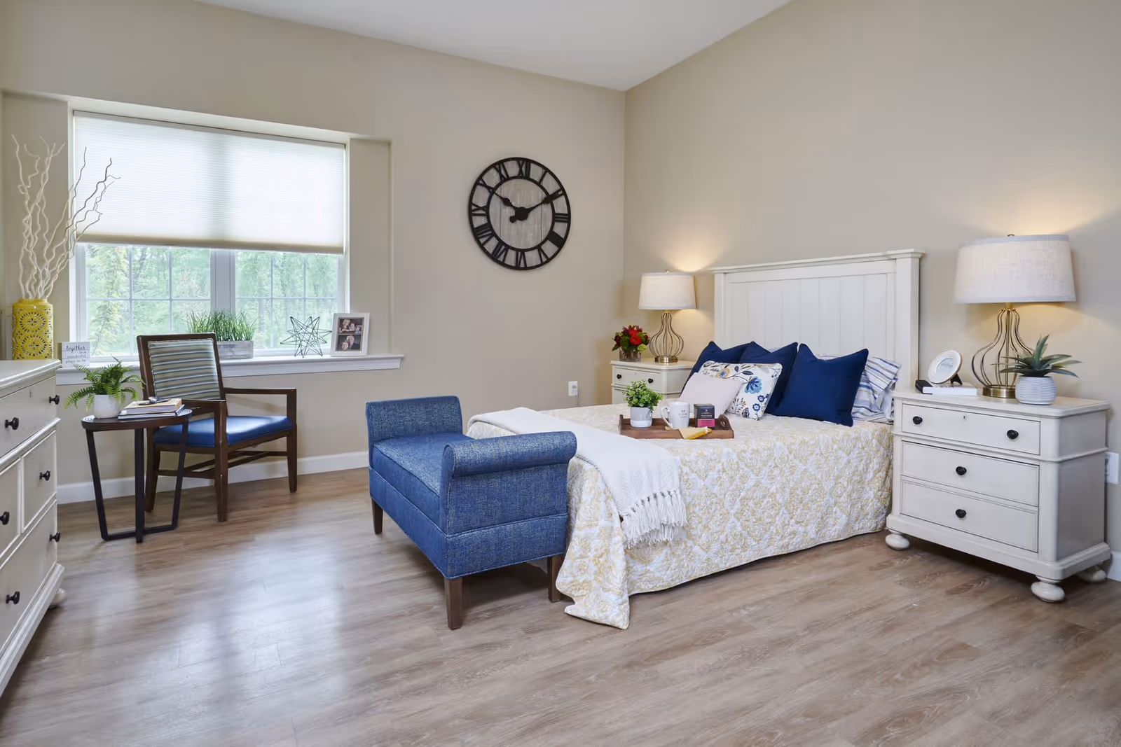 A bright and cozy bedroom with a large window covered by a white blind. The room features a white wooden bed with a yellow patterned bedspread, blue and white pillows, and a blue upholstered bench at the foot of the bed. There are two white nightstands with lamps on either side of the bed, a wooden chair with a blue cushion next to a small round table, and a white dresser. A large black wall clock hangs on the beige wall above the bed.