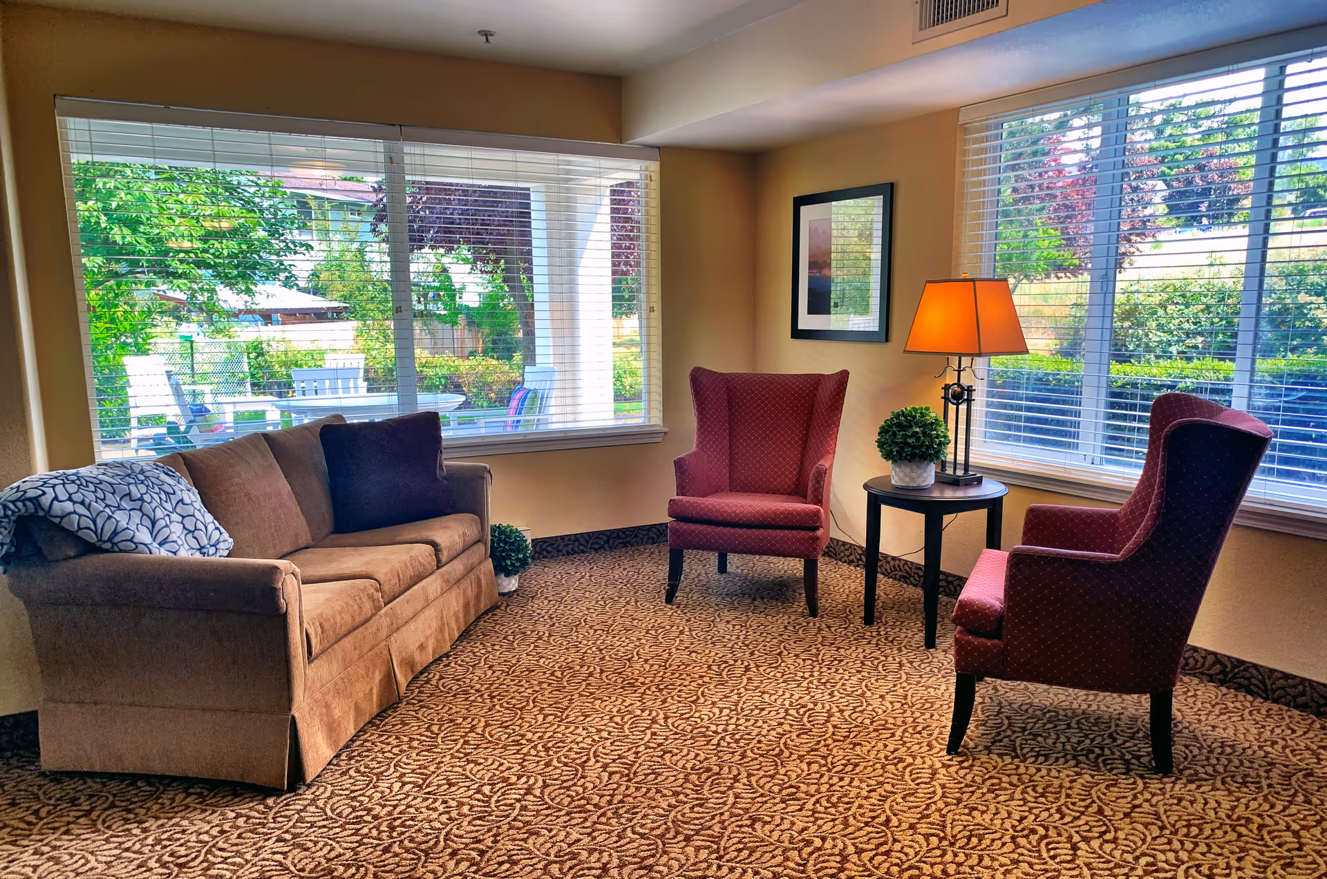 A cozy sitting area in a senior living facility with a brown sofa, two red armchairs, a small round table with a lamp and a potted plant, and large windows showing a green outdoor garden area.