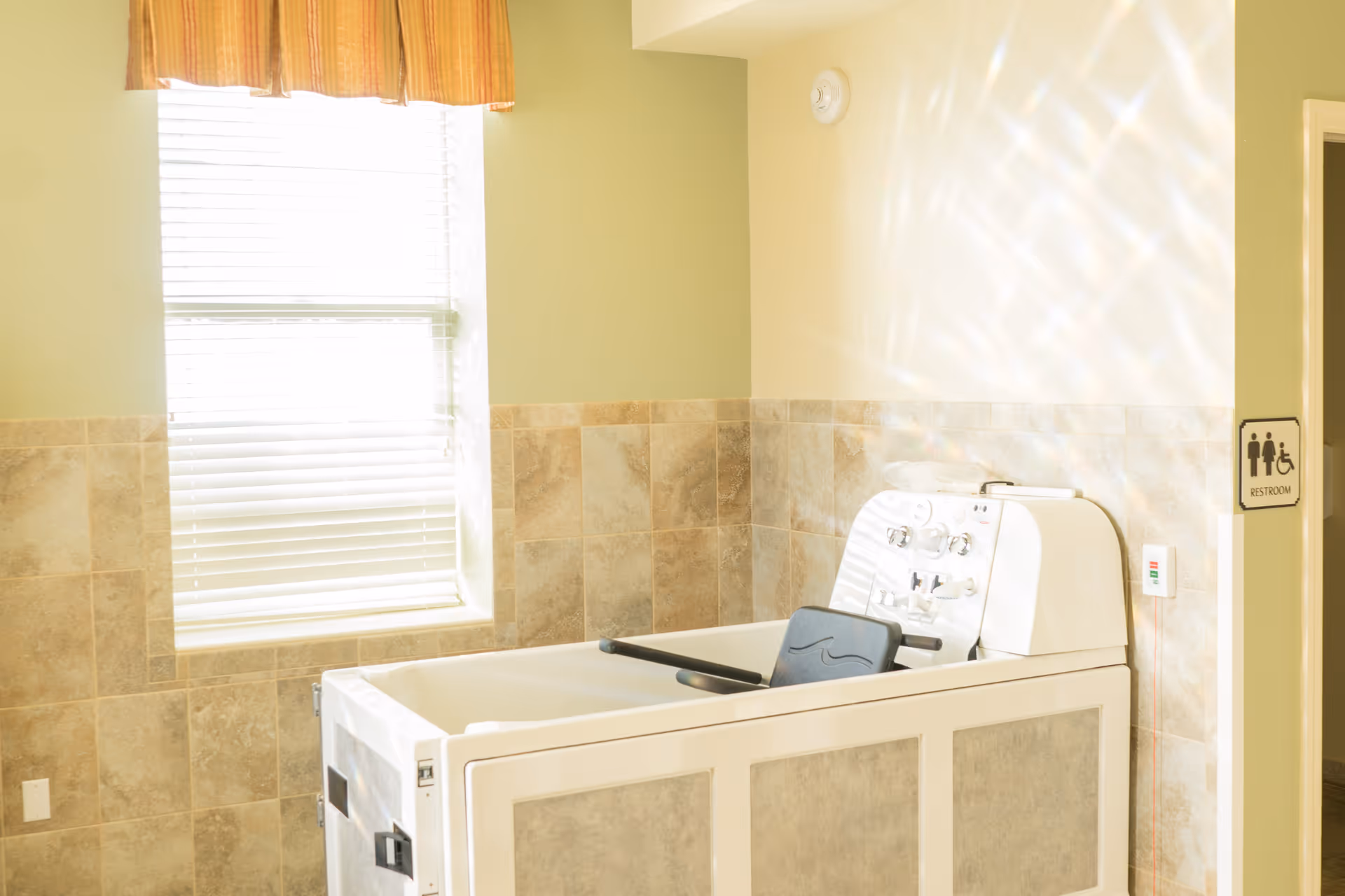 A bright bathroom with beige tiled walls and a large window with blinds and a valance. The room features a specialized walk-in bathtub with controls and a seat, designed for accessibility. A restroom sign indicating accessibility is visible on the wall near a doorway.