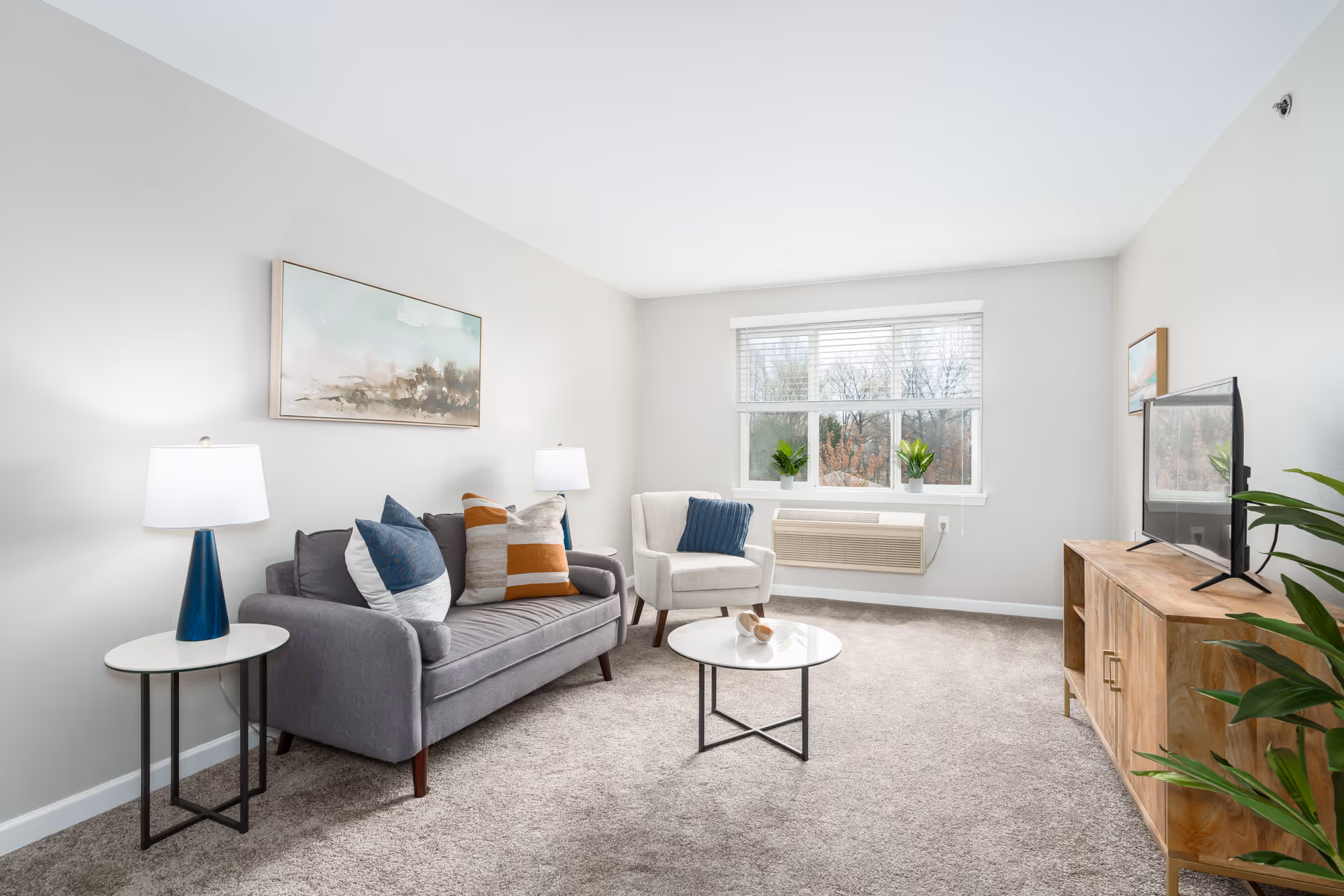 A bright living room with a gray sofa adorned with multiple cushions, a white armchair with a blue pillow, a round white coffee table, and a wooden TV stand with a flat-screen television. Two table lamps with white shades sit on either side of the sofa, and there are two small potted plants on the windowsill. The room has beige carpeting, light gray walls, and a large window with white blinds letting in natural light.