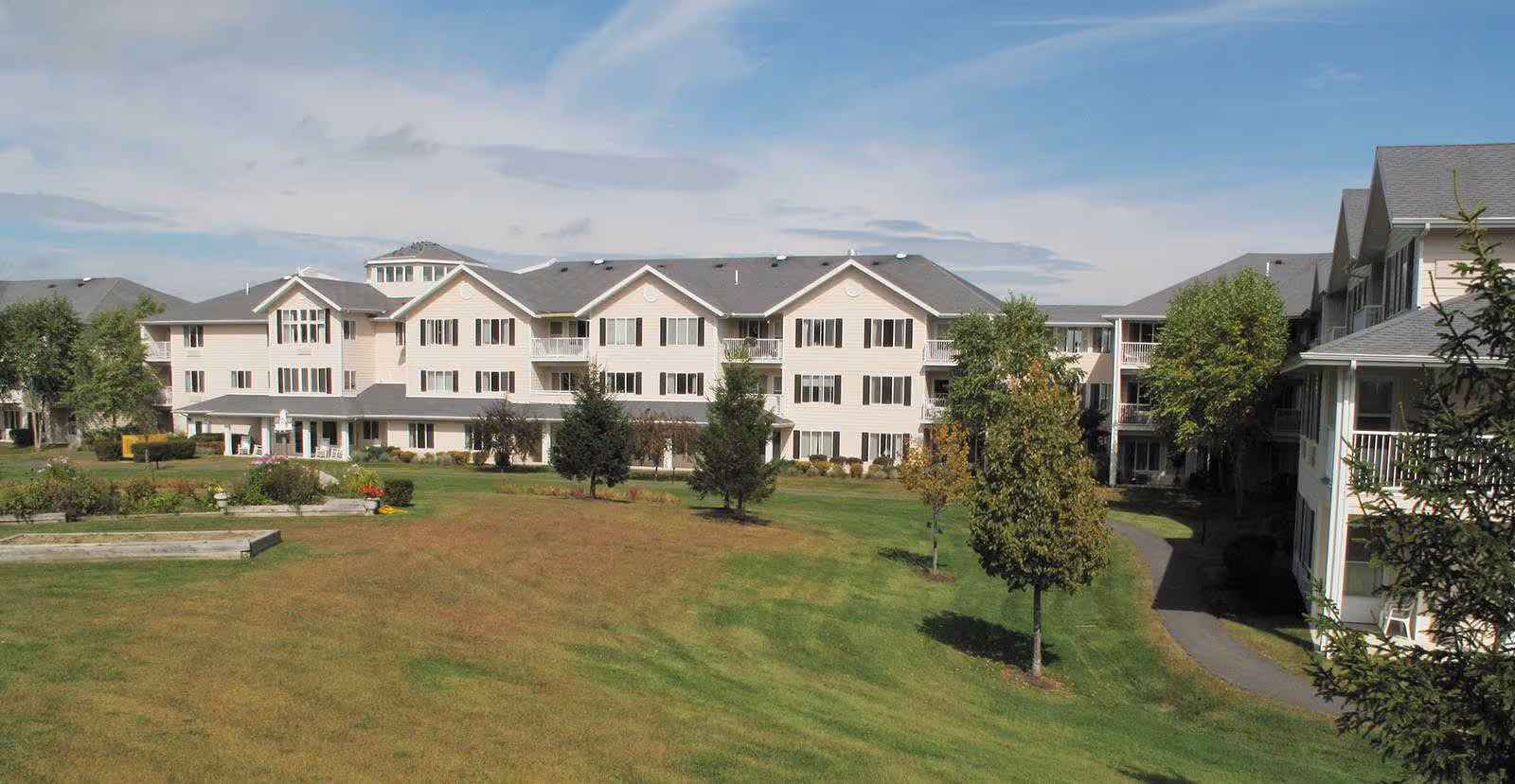 Exterior view of a large senior living facility building with multiple floors, beige siding, and dark shutters. The building is surrounded by a well-maintained lawn with several trees and a clear sky above.