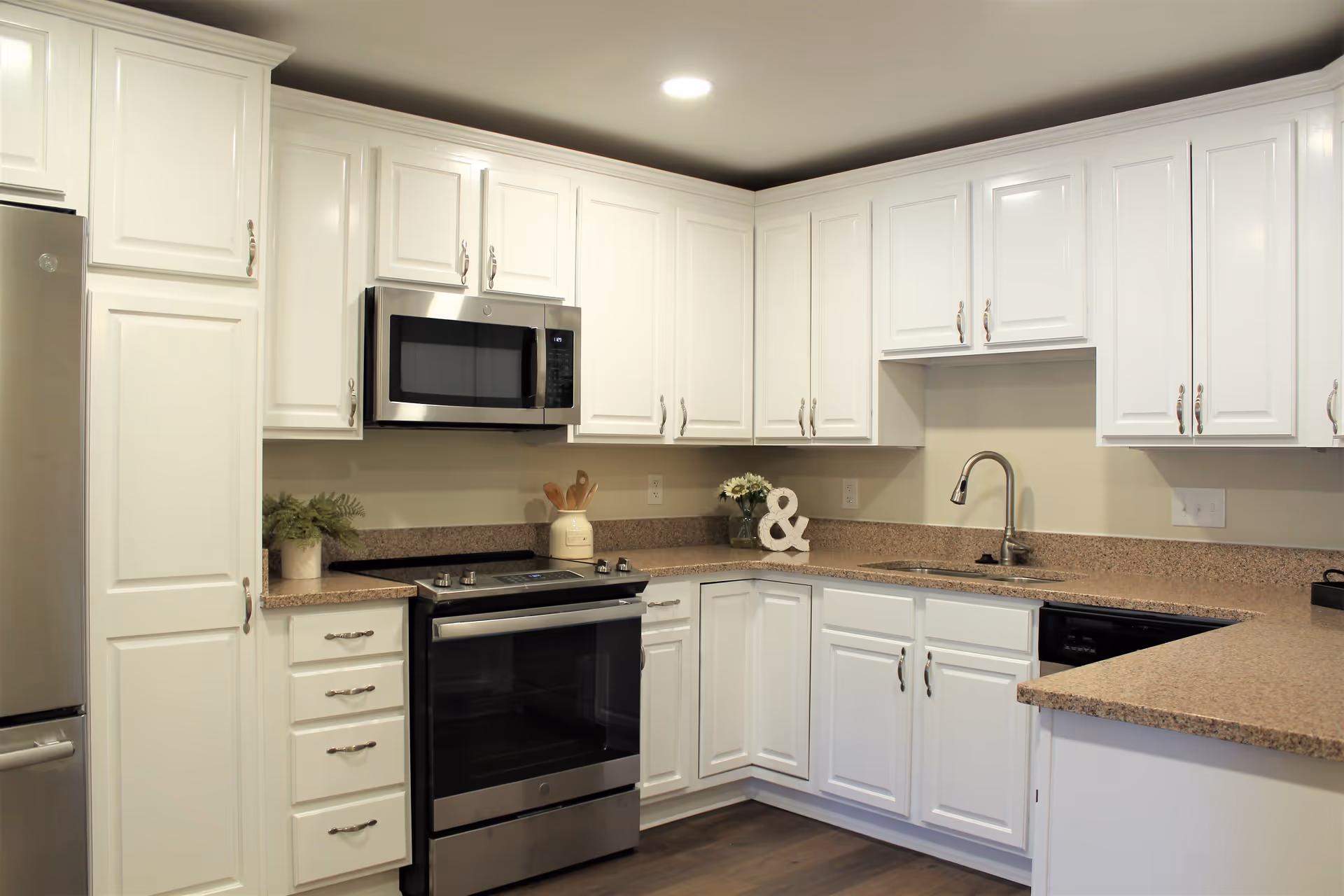 Modern kitchen with white cabinets, stainless steel appliances including a refrigerator, microwave, and oven, beige countertops, a sink with a faucet, and decorative items such as a plant, wooden utensils in a jar, flowers, and an ampersand symbol on the counter.