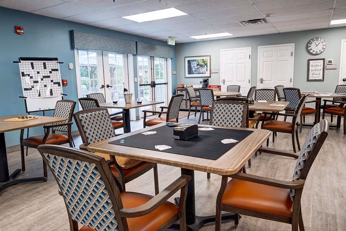 A well-lit activity room with multiple tables and chairs arranged for group games. One table in the foreground has playing cards and a card holder on a black mat. In the background, there is a large crossword puzzle board on an easel near glass doors leading outside. The walls are painted light blue and decorated with framed pictures and a clock.