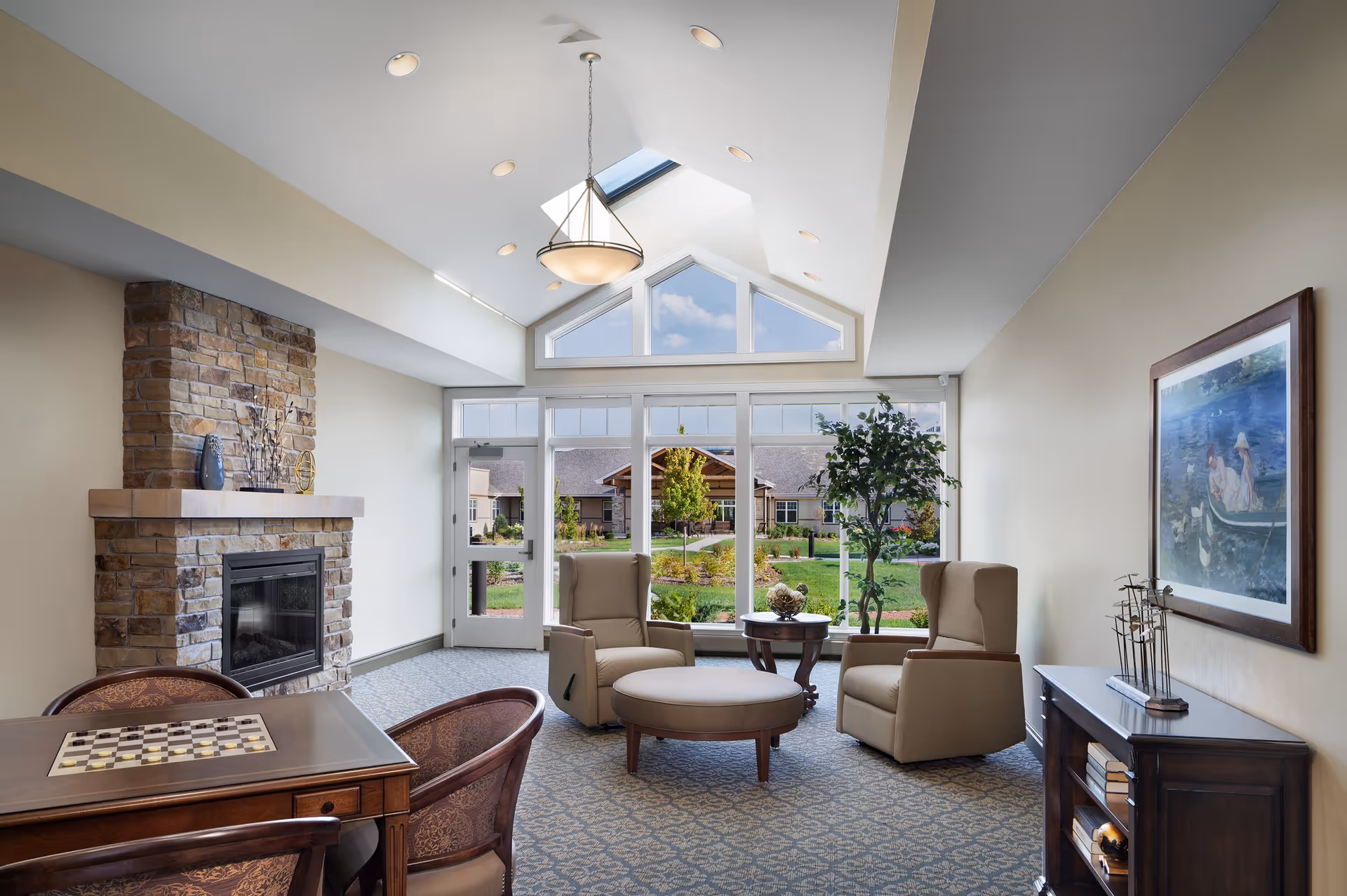 A bright and cozy living room area with two beige armchairs, a round ottoman, a small round wooden table, and a stone fireplace. Large windows and a glass door provide a view of a green garden and buildings outside. The room features a high ceiling with a skylight and a hanging light fixture. There is also a wooden table with a checkerboard and two chairs, a wooden cabinet with books, and a framed painting on the wall.