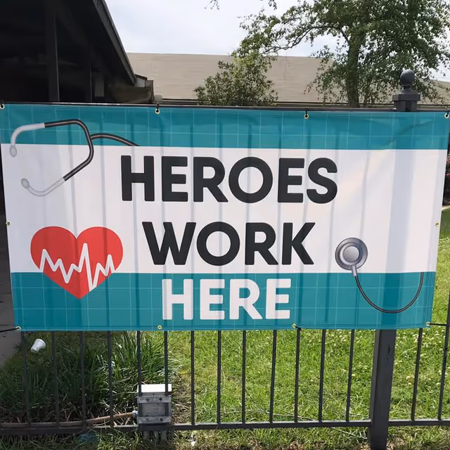 A banner hanging on a metal fence with the text 'HEROES WORK HERE' in bold letters. The banner features a red heart with a white heartbeat line on the left and a stethoscope graphic on both the left and right sides. The background of the banner is teal and white. Behind the fence, there is grass, some plants, and part of a building with trees visible.