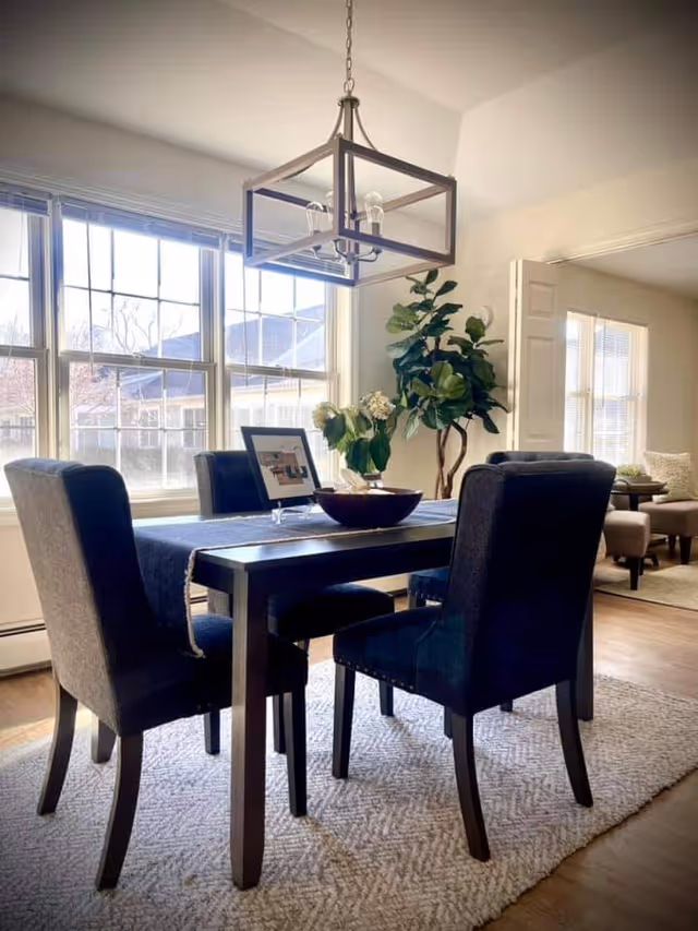 A bright dining area with a dark wooden table and four upholstered chairs on a light textured rug. A modern rectangular chandelier hangs above the table. There is a large window letting in natural light, a potted plant in the corner, and a glimpse of a living room with chairs and a small table in the background.