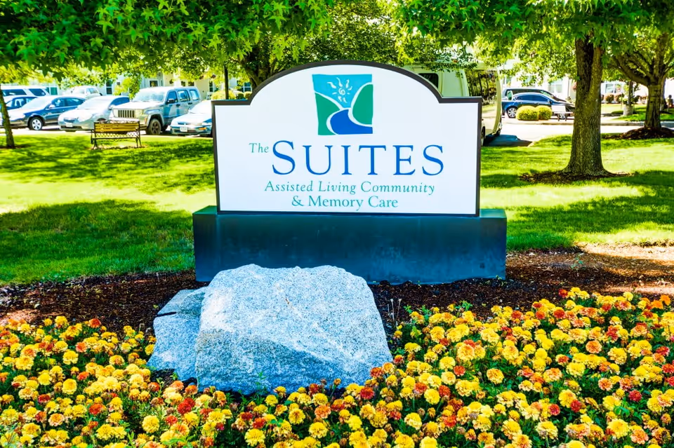 Outdoor view of a sign for The Suites Assisted Living Community & Memory Care, surrounded by a flower bed with yellow and orange flowers, green grass, trees, and parked cars in the background.
