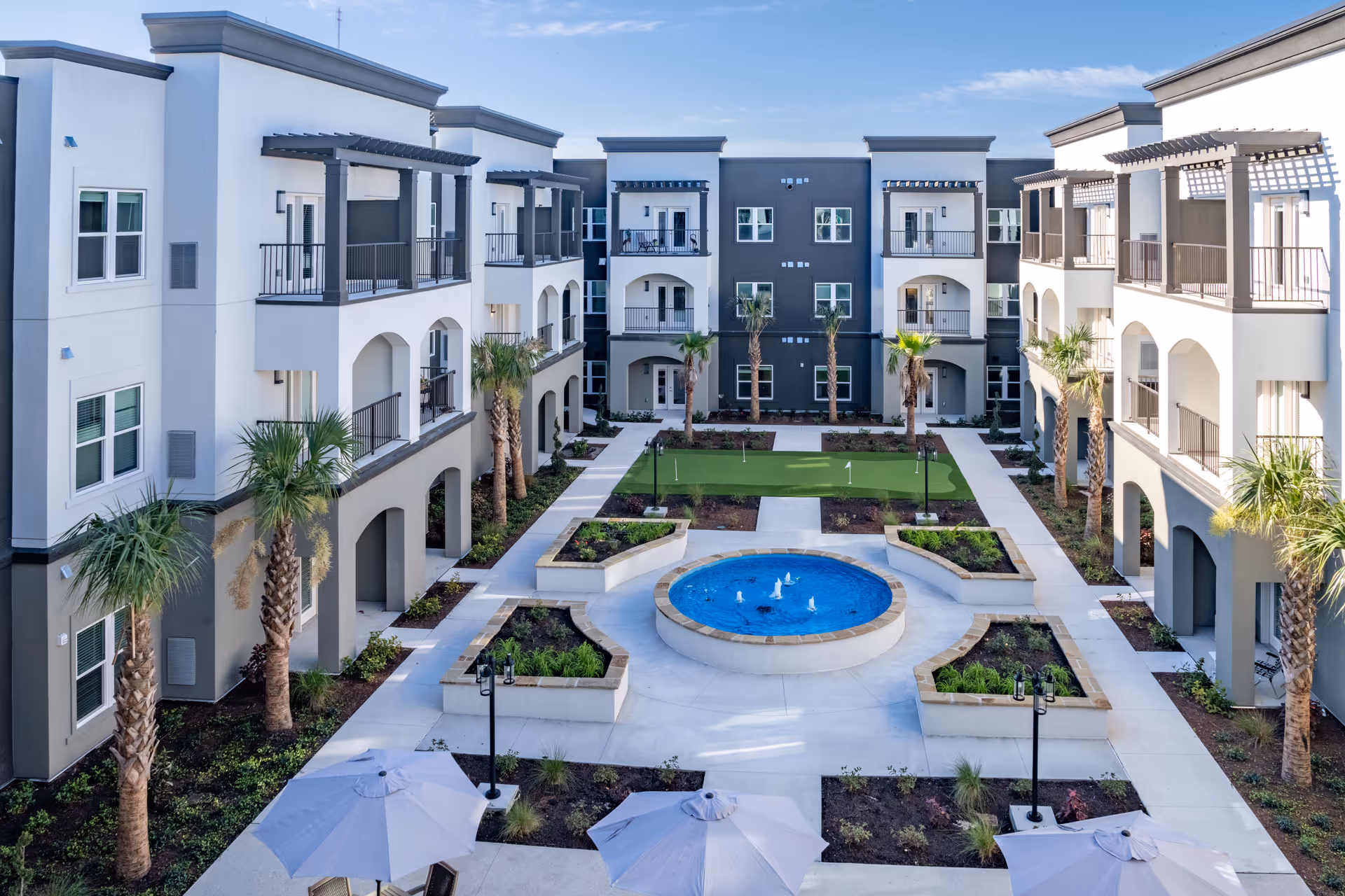 View of a modern senior living facility courtyard at The Goldton at Venice featuring a central circular water fountain, landscaped garden beds, palm trees, a putting green, and surrounding three-story buildings with balconies and shaded walkways under a clear blue sky.