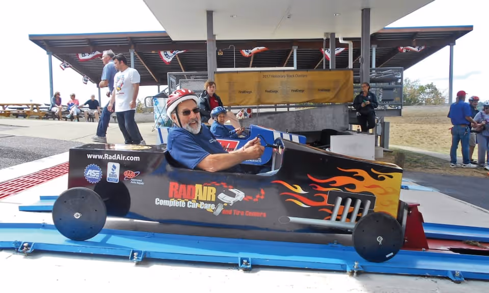 An elderly man wearing a red helmet and sunglasses is sitting in a black soapbox derby car with flame decals and the logo 'RadAir Complete Car Care and Tire Centers' on the side. He is smiling and holding the steering wheel. Behind him, a young boy wearing a blue helmet is seated in another soapbox car. Several people are standing and sitting in the background near a covered pavilion with picnic tables and a yellow banner.