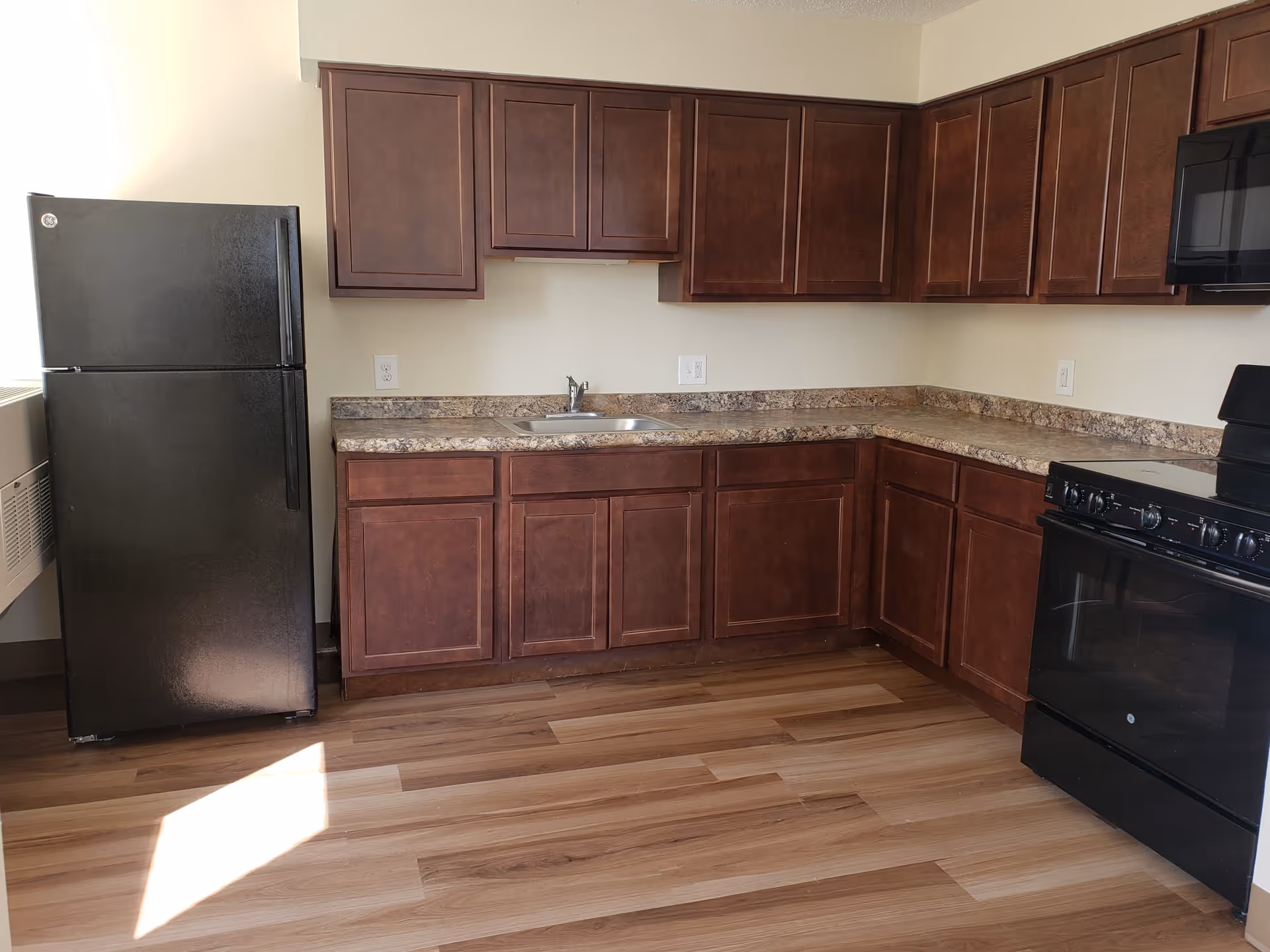 A kitchen with dark wood cabinets, a black refrigerator, a black stove with oven, a microwave, a stainless steel sink, and a countertop with a marble pattern. The floor is wooden with a light and dark brown pattern.