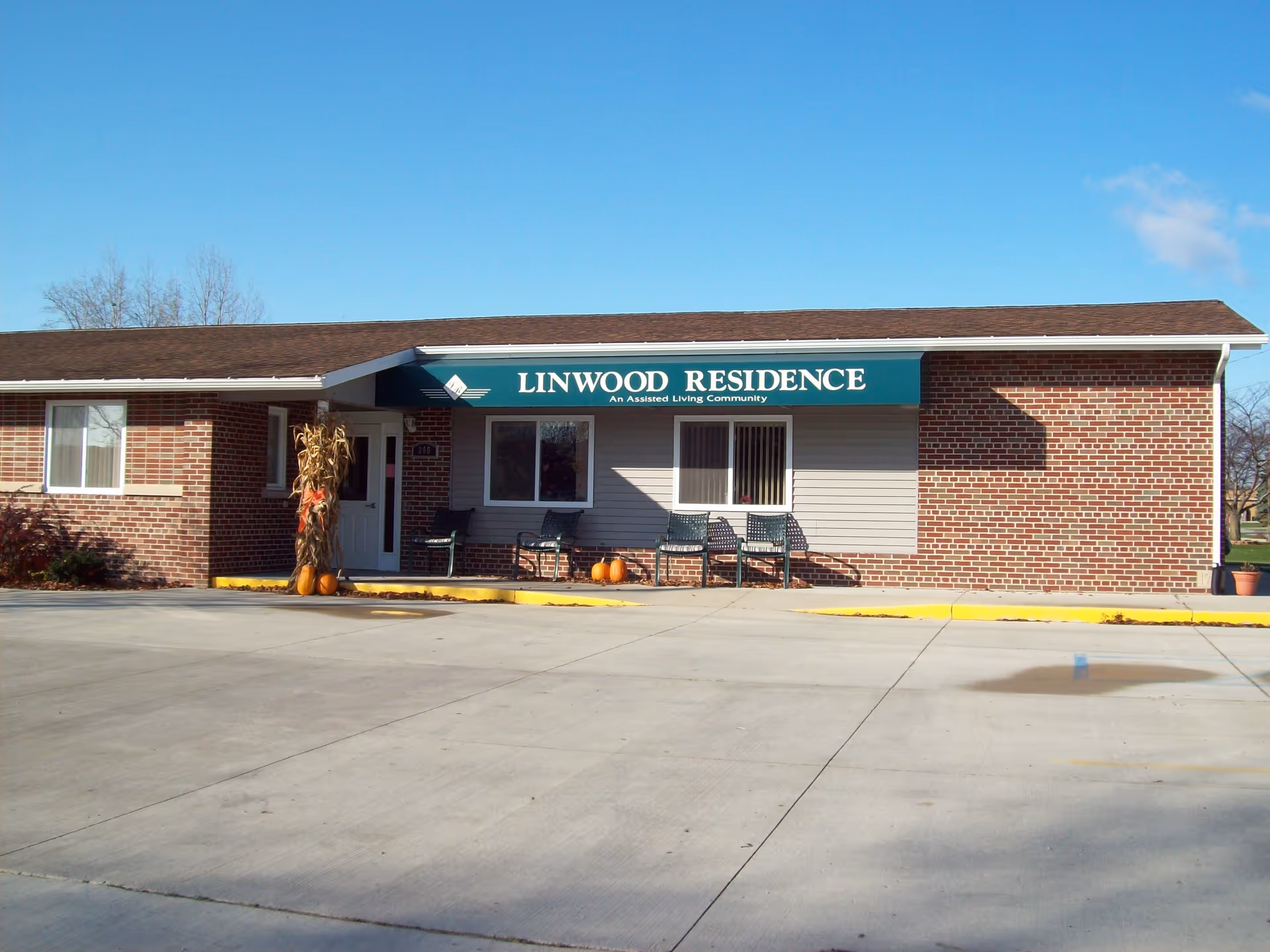 Exterior view of Linwood Residence, an assisted living community, showing a single-story brick building with a green awning above the entrance. There are chairs and pumpkins placed outside near the entrance, and the sky is clear and blue.