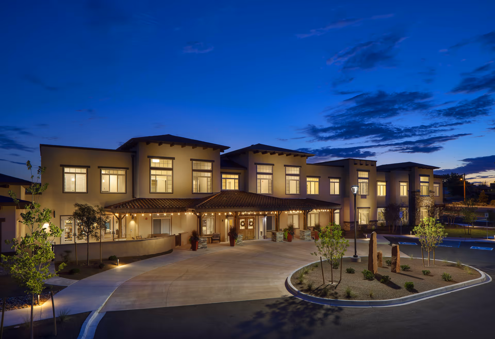 Exterior view of MorningStar Assisted Living & Memory Care of Rio Rancho building at dusk with lights on inside and a curved driveway in front, surrounded by small trees and landscaping.