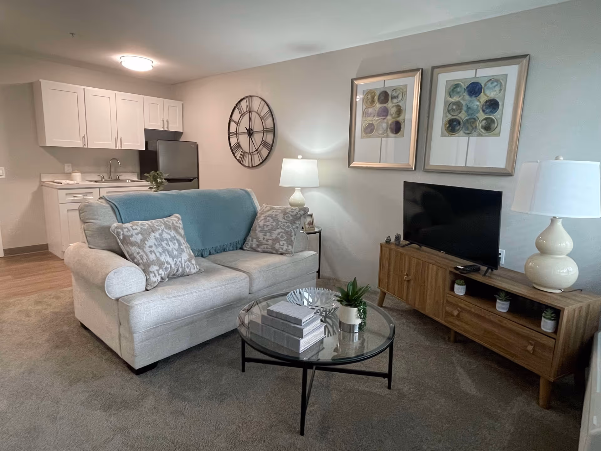 A cozy living room area with a beige sofa adorned with patterned pillows and a blue throw blanket. In front of the sofa is a round glass coffee table with decorative items including books and small plants. To the right, there is a wooden TV stand with a flat-screen TV, two table lamps, and small potted plants. The wall behind the TV has two framed abstract art pieces and a large decorative wall clock. In the background, a small kitchenette with white cabinets, a sink, and a refrigerator is visible.