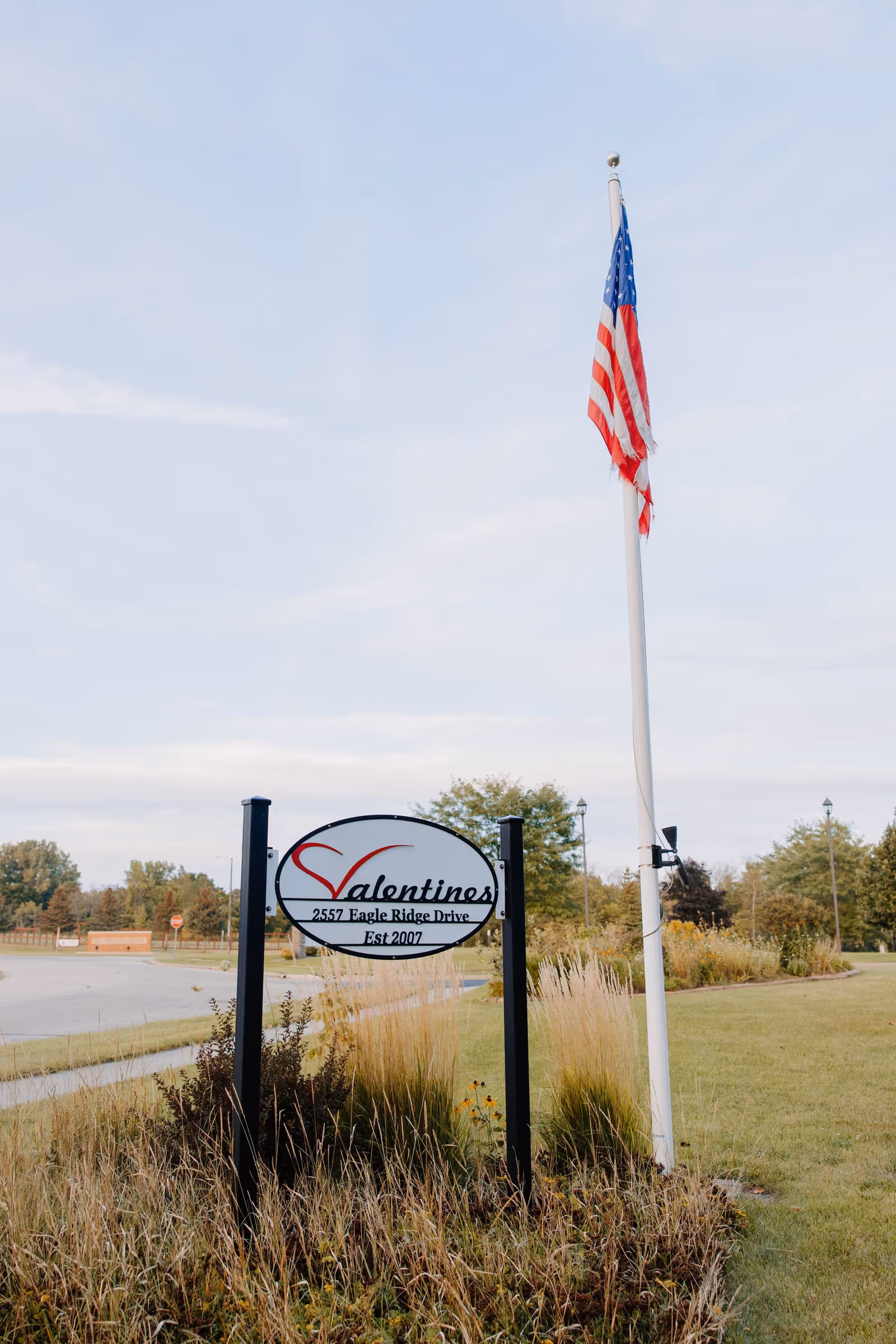 Outdoor view of a sign for Valentines Assisted Living located at 2557 Eagle Ridge Drive, established in 2007, with an American flag on a flagpole nearby and grassy landscaping around the sign.