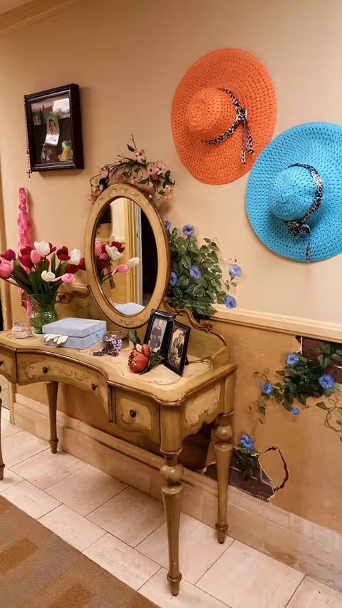 A vintage-style wooden console table with an oval mirror mounted on the wall above it. The table holds a vase of tulips, a blue box, framed photos, and decorative items. Two large woven hats, one orange and one blue, hang on the wall. The wall is decorated with painted floral designs and a small framed picture.