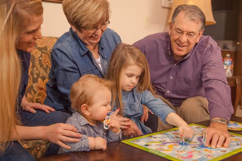 Two elderly adults and two young children sitting around a wooden table playing a colorful board game together in a cozy living room setting.