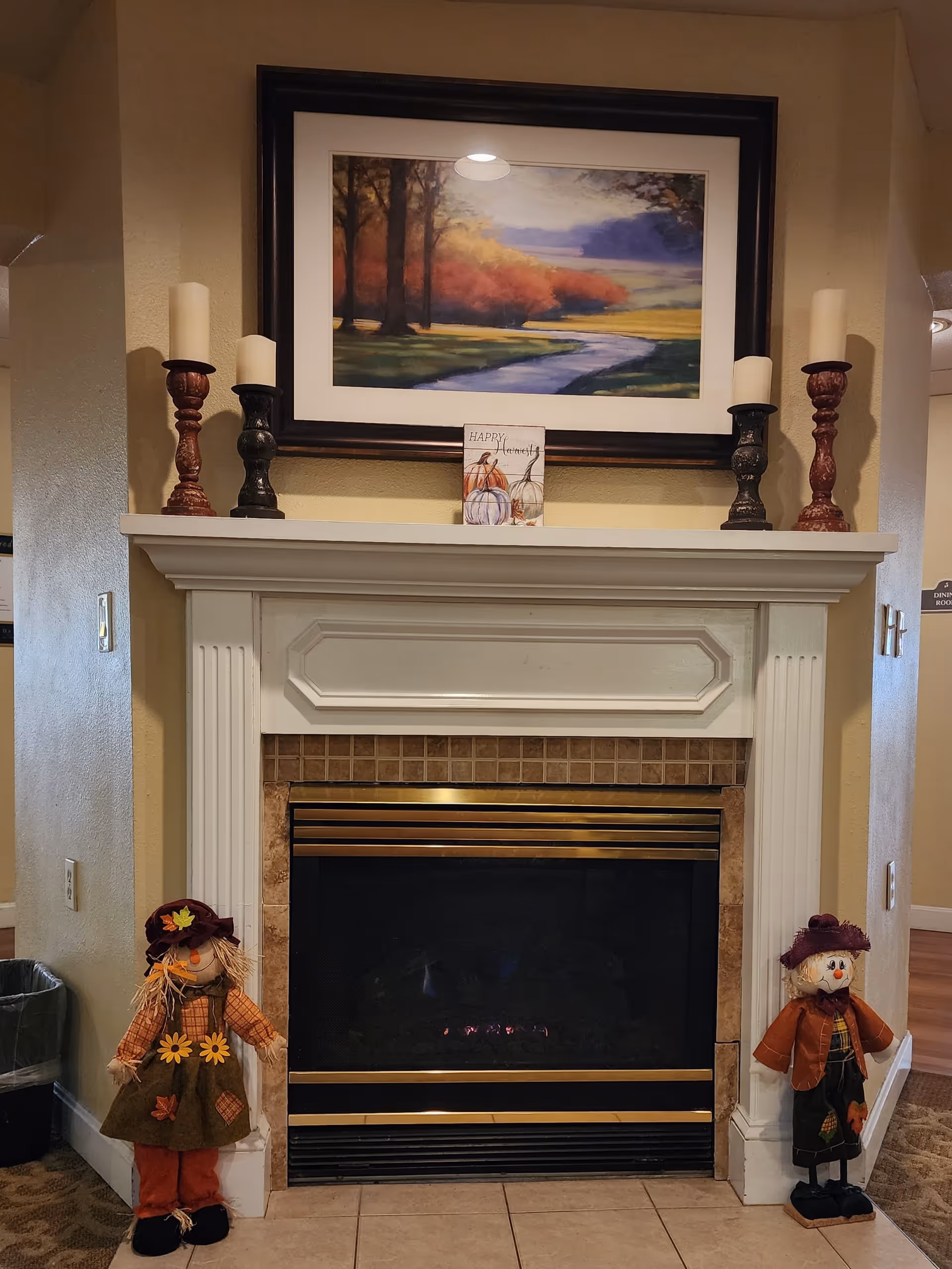 A cozy indoor fireplace with a white mantel decorated with four candles on holders and a framed landscape painting above it. Two scarecrow dolls dressed in autumn-themed clothing stand on either side of the fireplace. The floor is tiled and the walls are painted beige.