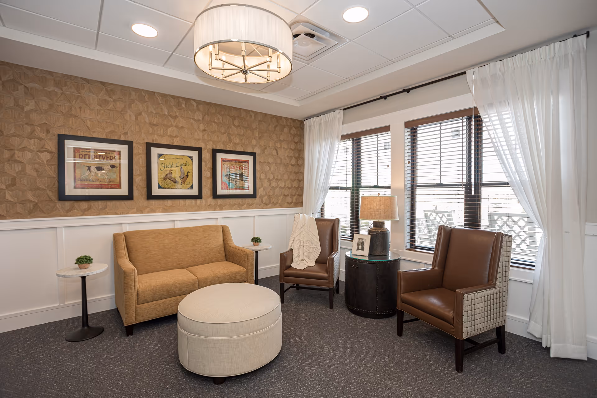 A cozy living room area with a beige loveseat, two brown armchairs, a round beige ottoman, and two small side tables with plants. The room has a textured tan accent wall with three framed pictures, large windows with white curtains and blinds, and a ceiling light fixture.
