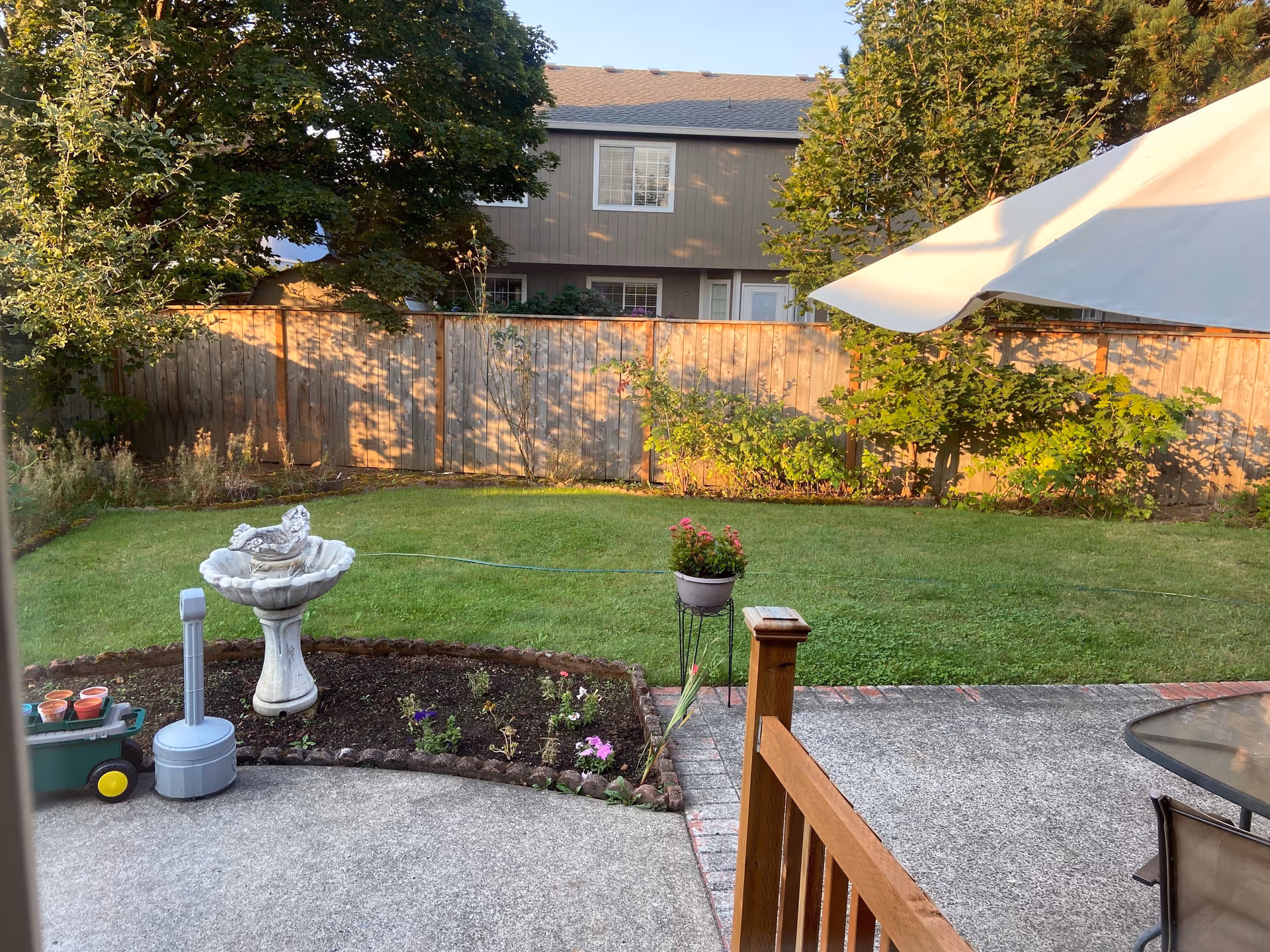 Backyard patio and lawn with a garden bed, birdbath, potted plants, umbrella and a wooden fence with a neighboring house beyond.