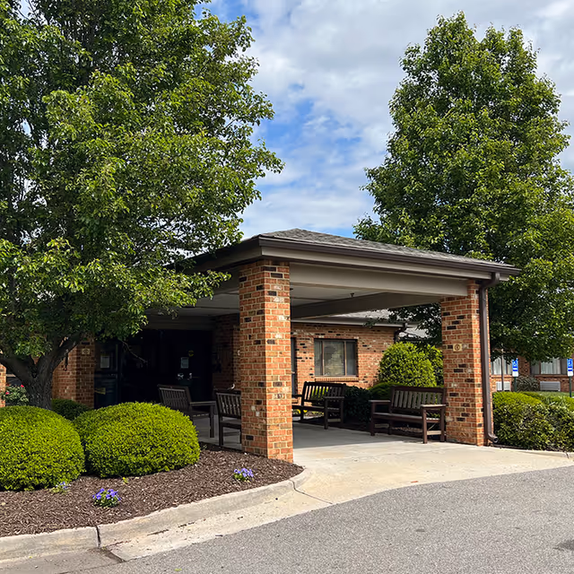 Entrance area of a brick building with a covered porch supported by brick columns. There are several wooden benches under the porch. The surrounding area has well-maintained bushes, trees, and a paved driveway. The sky is partly cloudy.