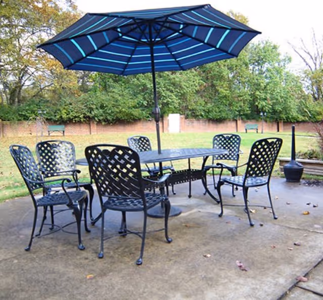 Outdoor patio area with a black metal table and six matching chairs arranged around it. A large blue umbrella with light blue stripes is open above the table. The patio is surrounded by grass, trees, and a brick wall in the background.