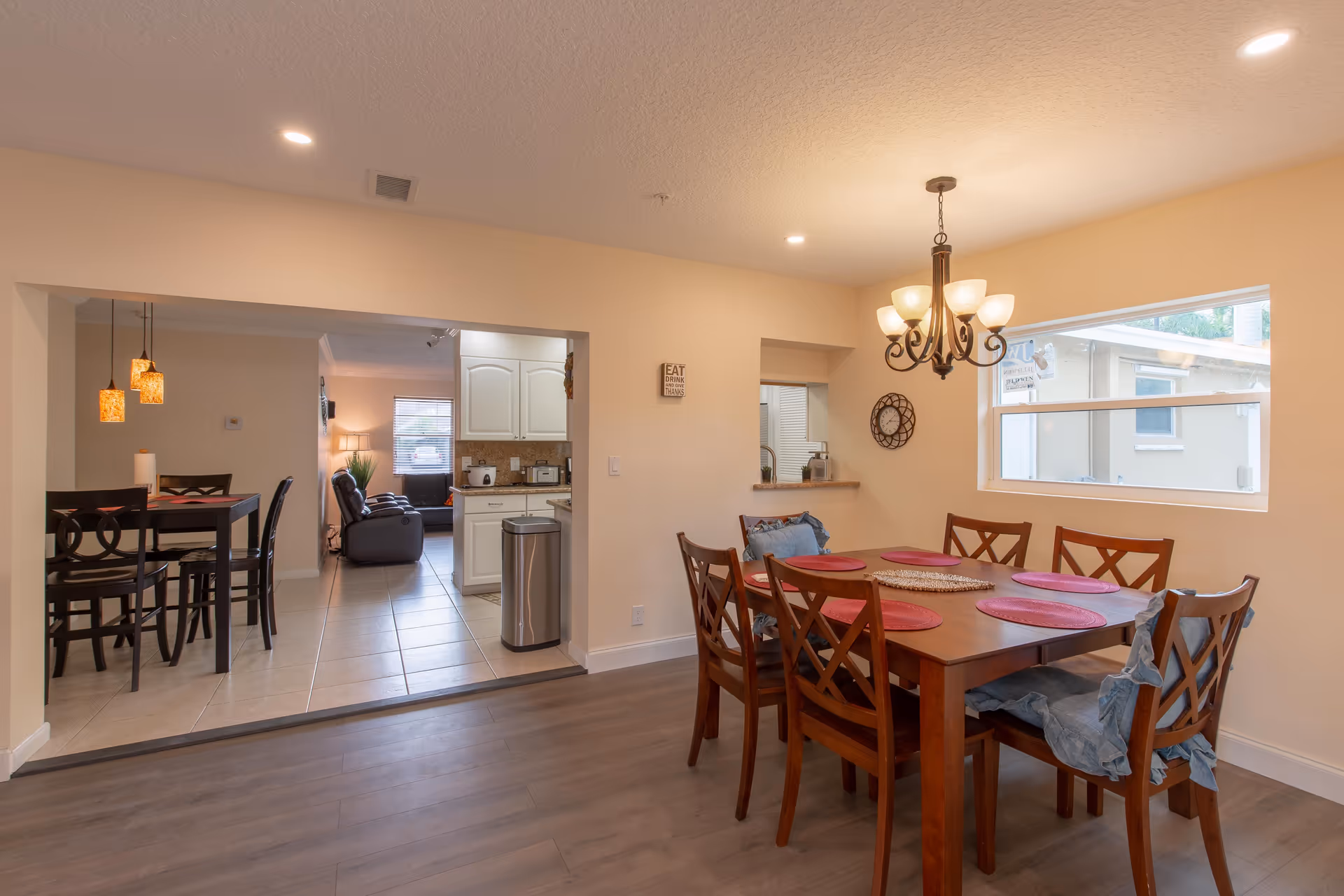 Interior view of a dining area in a Mediterranean Assisted Living Facility featuring a wooden dining table with six chairs, red placemats, and a chandelier overhead. The room has light-colored walls, a window letting in natural light, and an open doorway leading to a kitchen and living room area with additional seating and lighting fixtures.