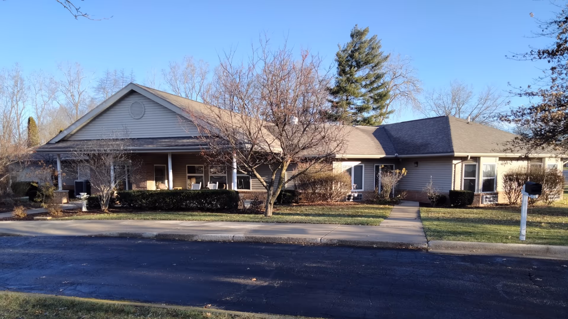 Exterior view of a single-story residential building with a covered porch, several windows, and a sidewalk leading to the entrance. The building is surrounded by leafless trees, bushes, and a grassy lawn under a clear blue sky.