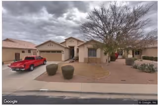 Exterior view of a single-story residential building with a driveway, a red pickup truck parked in front, desert landscaping with bushes and a tree, under a cloudy sky.