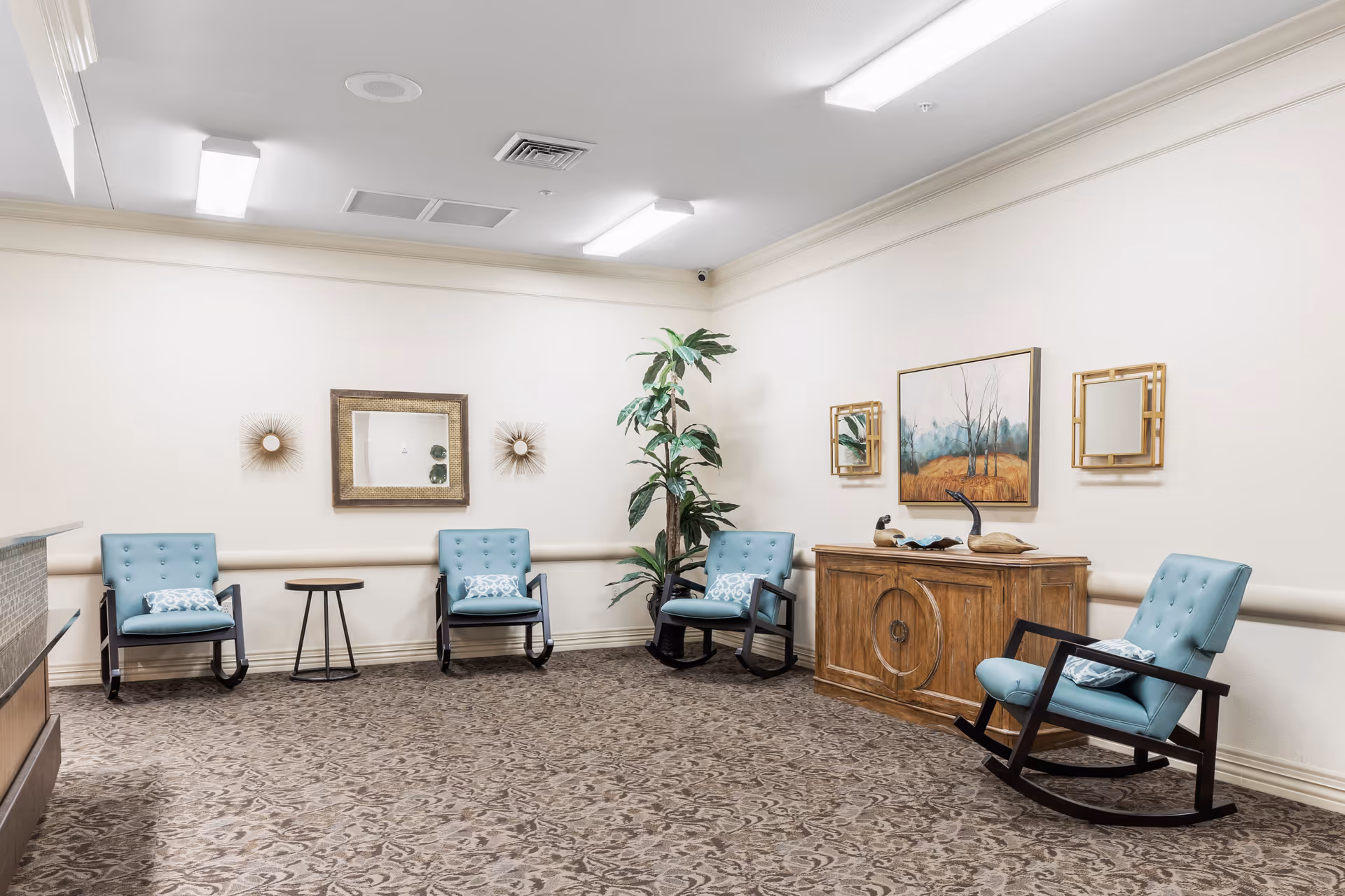 Bright seating area with blue rocking chairs, side tables, a wooden cabinet, artwork, and a potted plant in a common room.