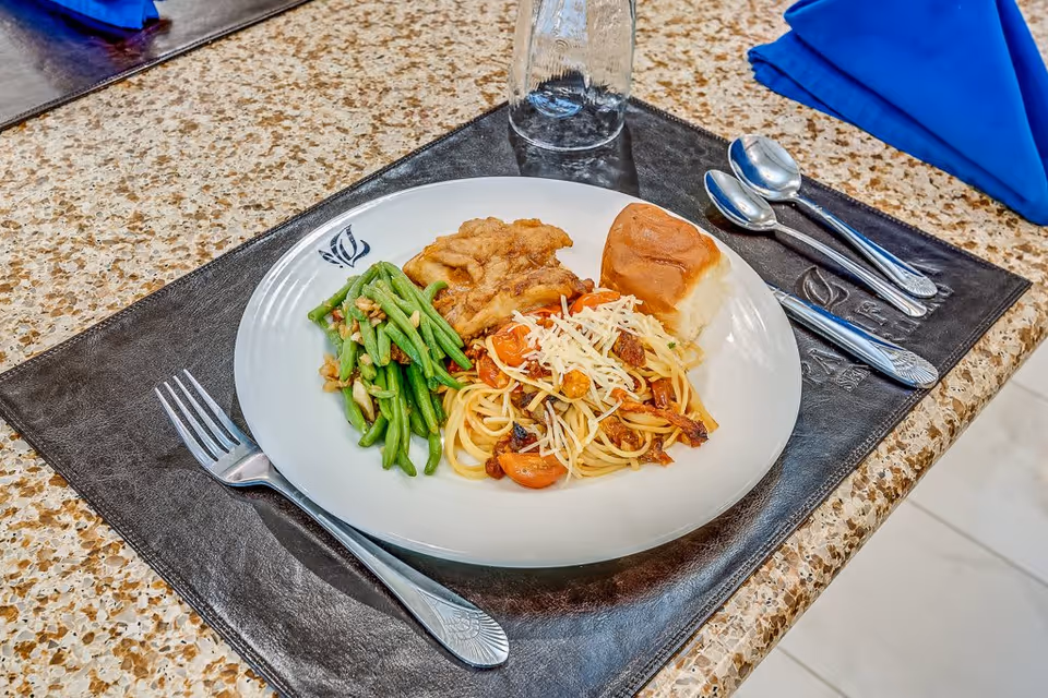 A plate of food on a placemat with a fork, two spoons, and a glass. The plate contains green beans with walnuts, spaghetti topped with shredded cheese and cherry tomatoes, a bread roll, and a piece of fried chicken. The setting is on a speckled countertop with a folded blue napkin in the background.
