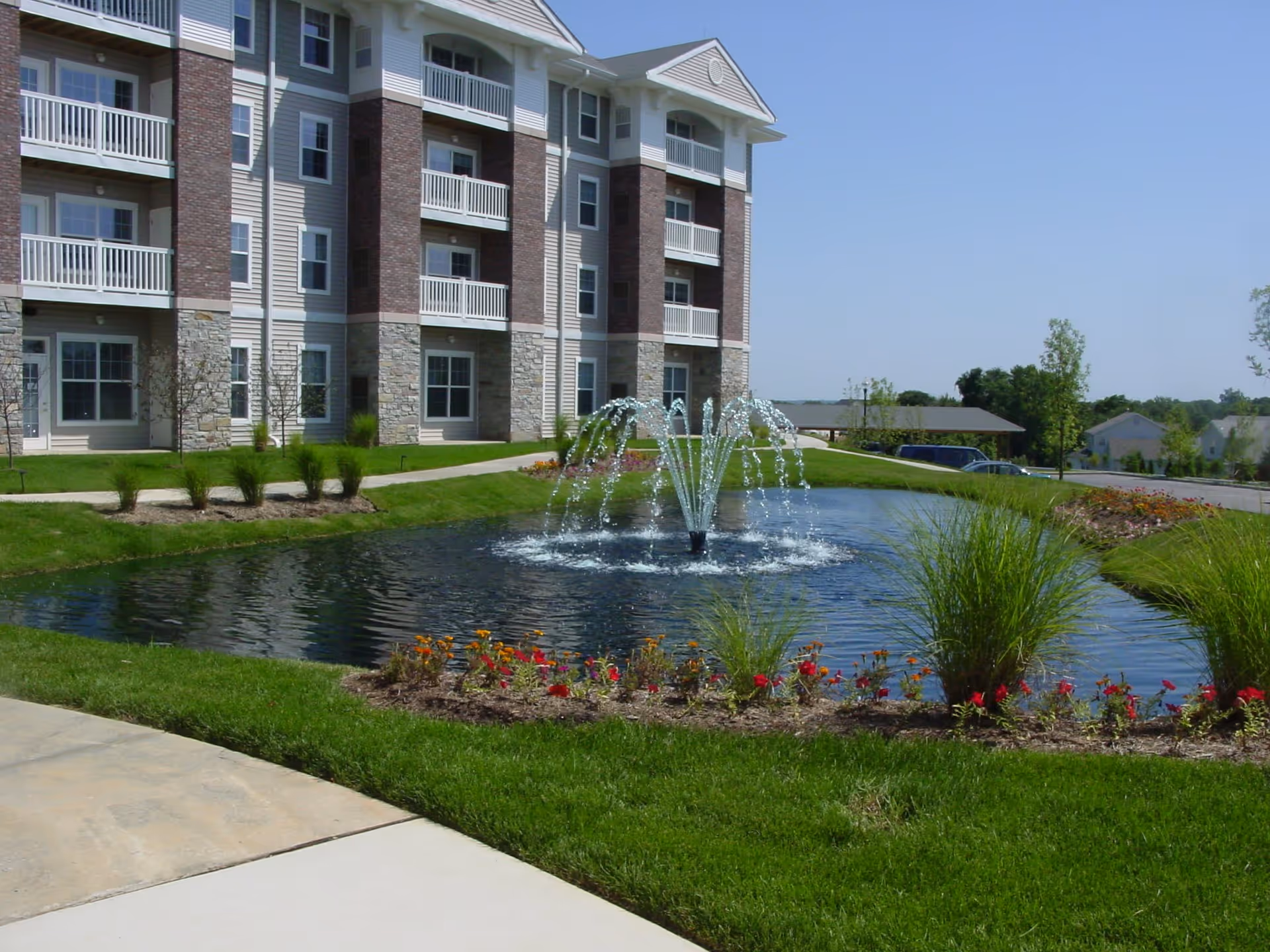 Outdoor view of a senior living facility with a multi-story building featuring balconies. In front of the building is a landscaped area with a pond and a water fountain, surrounded by grass, flowers, and shrubs under a clear blue sky.