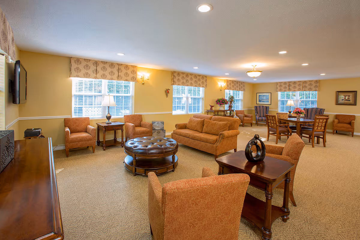 A spacious and well-lit living room area in a senior living facility with multiple seating arrangements including sofas, armchairs, and wooden tables. The room features beige walls with white trim, patterned valances on the windows, and carpeted flooring. Decorative elements include table lamps, framed artwork, and floral arrangements.
