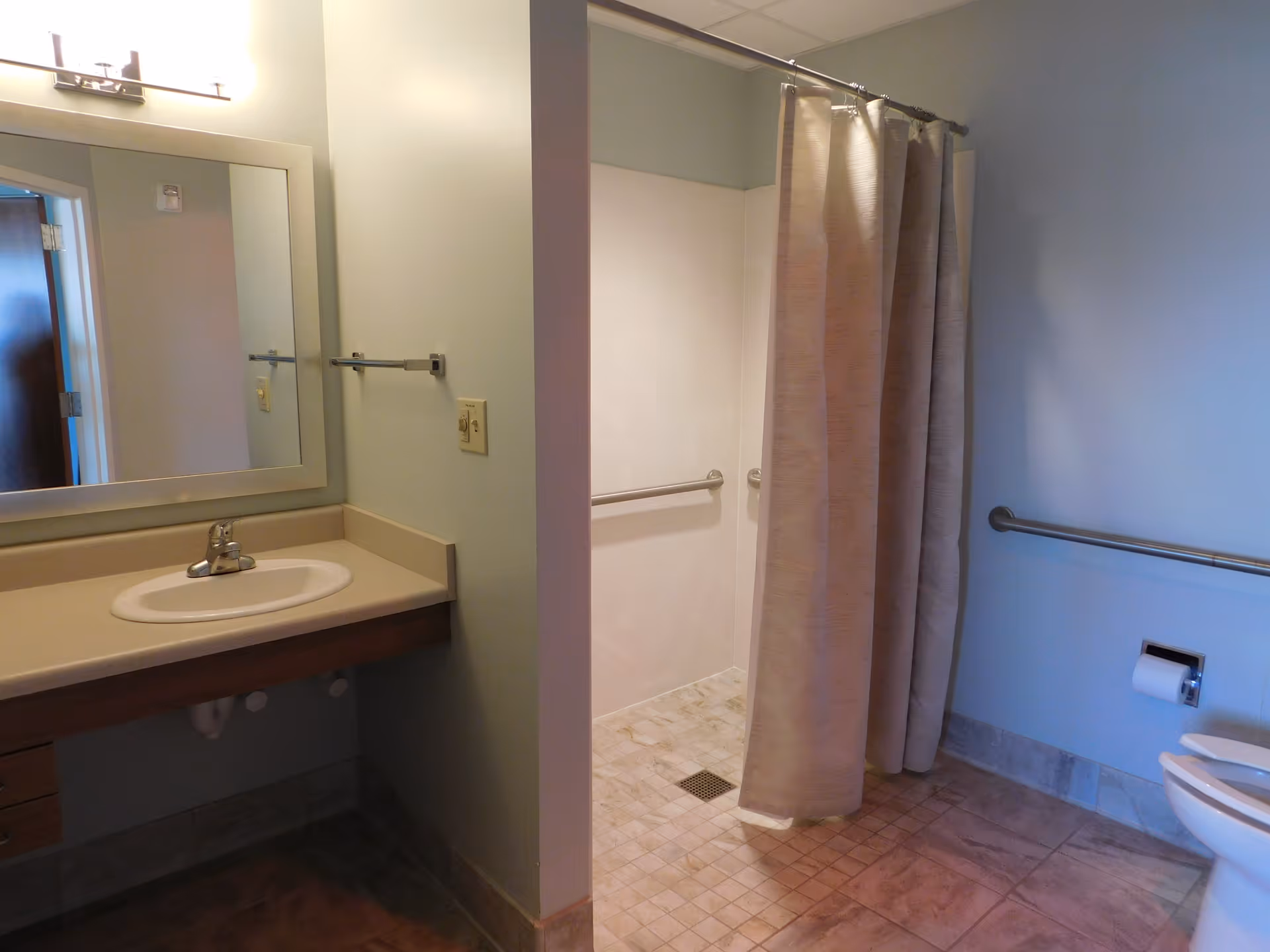 A bathroom with a sink and countertop on the left, a large mirror above the sink, a walk-in shower with a curtain in the center, and a toilet with a grab bar and toilet paper holder on the right. The floor is tiled and the walls are painted light blue.