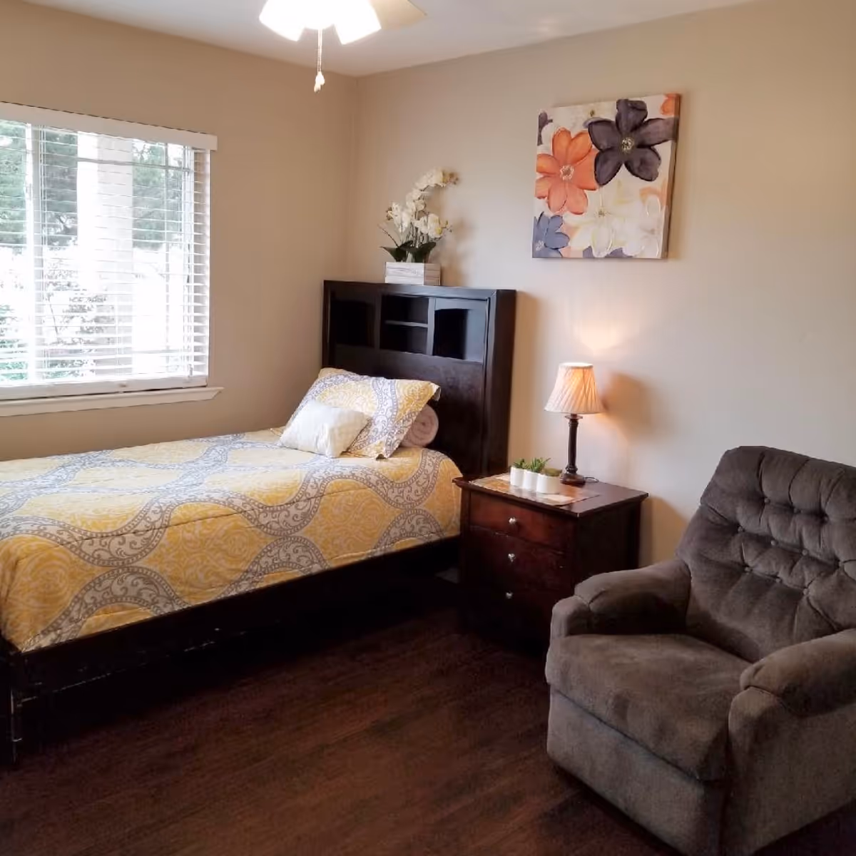 A cozy bedroom with a single bed covered in a yellow and gray patterned bedspread, a dark wooden headboard with a white flower arrangement on top, a wooden nightstand with a small lamp and potted plants, a comfortable gray armchair, and a window with white blinds letting in natural light.