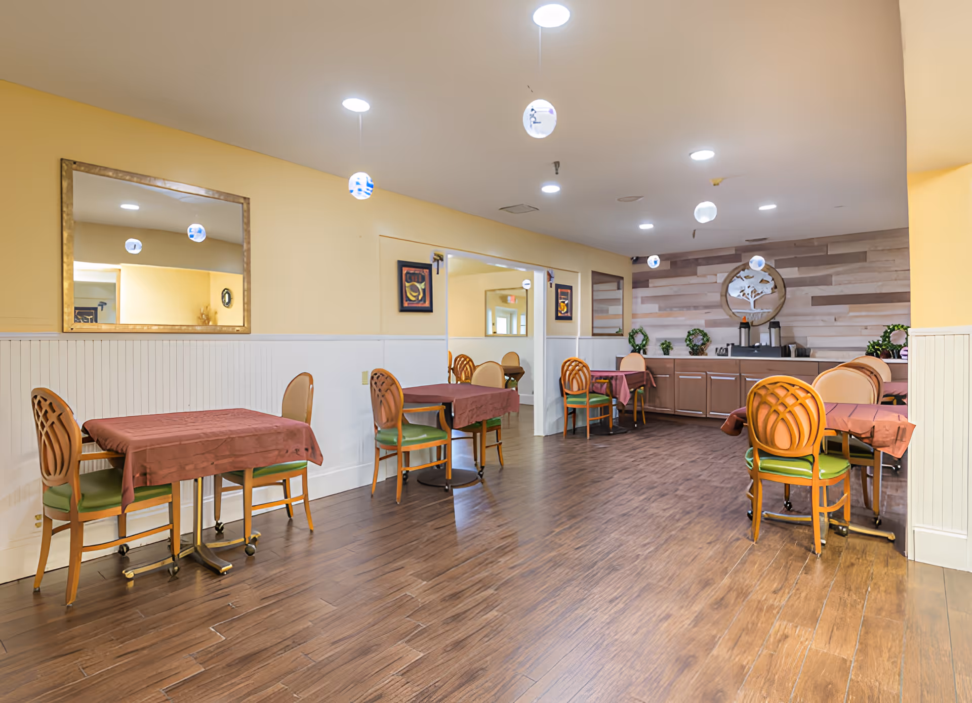 A dining room in Colonial Assisted Living at Miami with several tables covered with brown tablecloths and wooden chairs with green cushions. The room has yellow walls with white wainscoting, a large mirror on one wall, and a wooden accent wall with decorative wreaths and a circular wall art piece. The floor is wood, and the ceiling has recessed lighting.