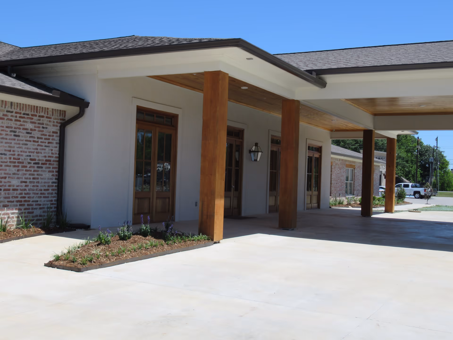 Exterior view of the entrance area of BeeHive Homes of Youngsville featuring a covered driveway with wooden support columns, glass-paneled wooden doors, brick and stucco walls, and a small landscaped garden bed with plants and flowers.