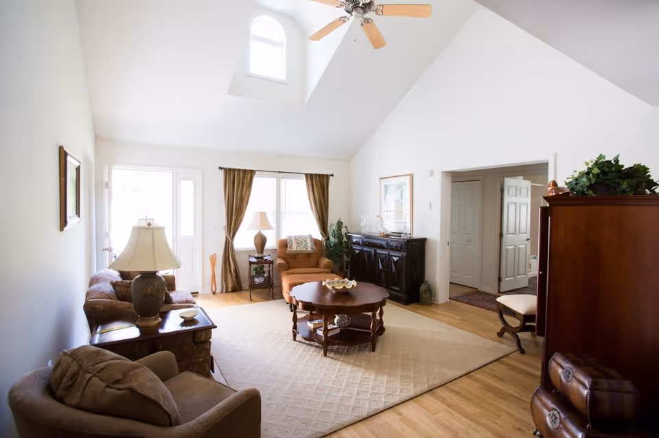 A bright living room with high vaulted ceilings and a ceiling fan. The room features a large window with brown curtains, a brown armchair, a matching sofa, a wooden coffee table on a beige rug, and a dark wooden cabinet. There are lamps on side tables and some decorative plants and artwork on the walls.