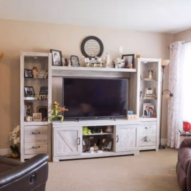 A cozy living room with a large flat-screen TV mounted on a white wooden entertainment center. The entertainment center has shelves and cabinets displaying framed photos, decorative items, and plants. There is a floor lamp and a window with light curtains on the right side, and part of a brown leather chair is visible in the foreground.