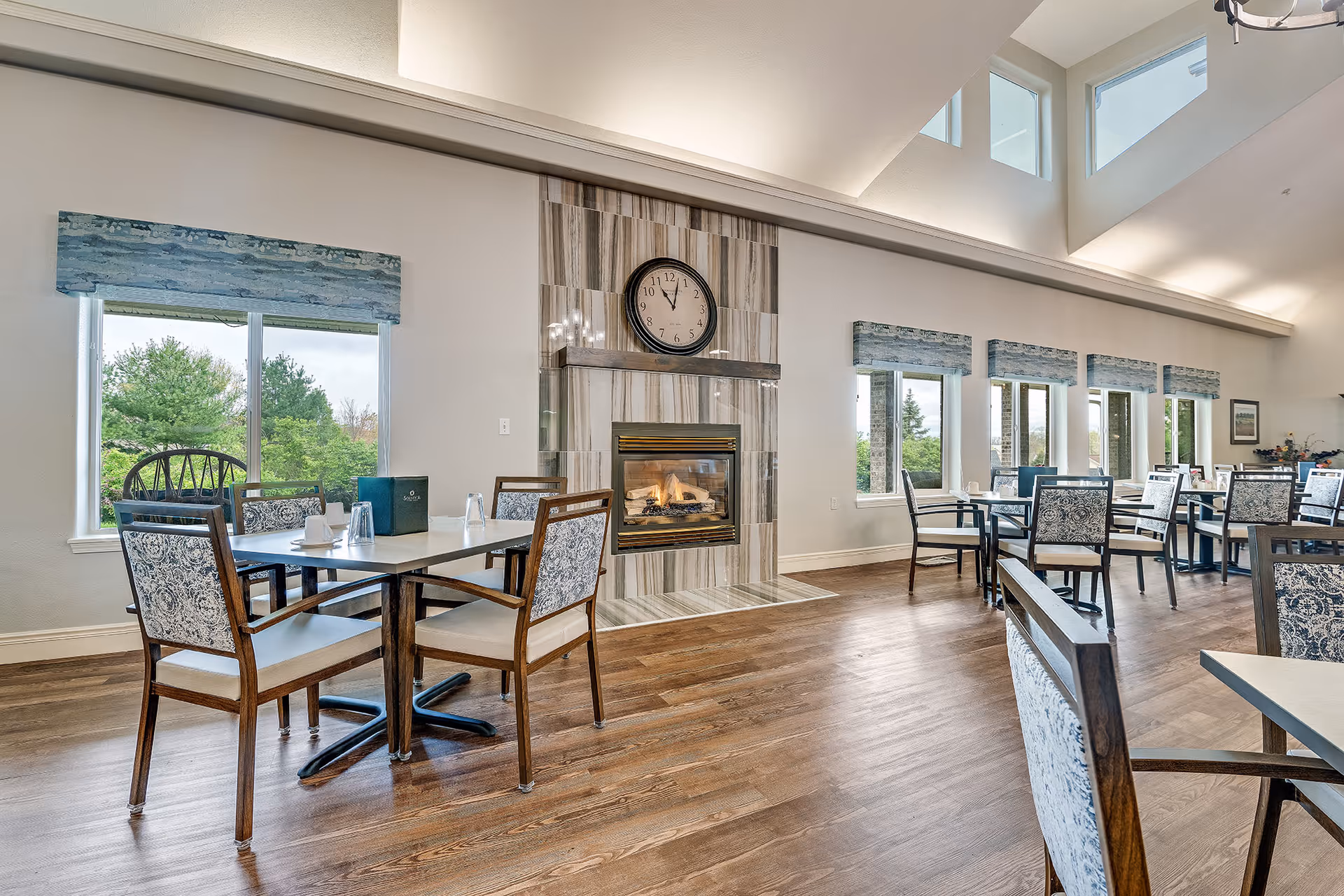 A bright dining room with multiple tables and chairs featuring patterned upholstery. A modern fireplace with a clock above it is centered on one wall. Large windows with blue valances let in natural light and offer views of greenery outside. The room has high ceilings with additional windows near the top.