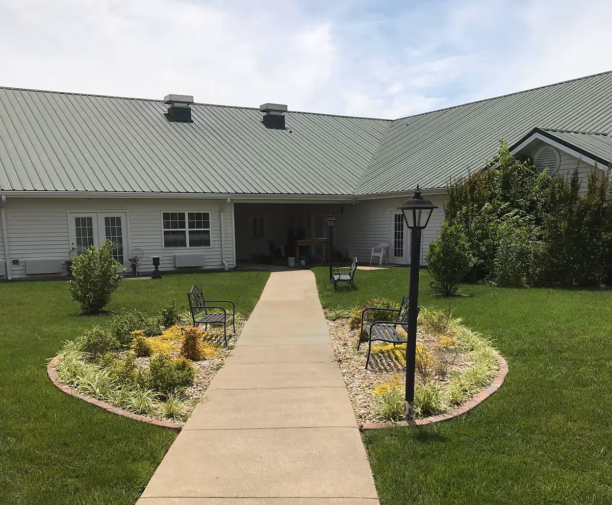 Outdoor courtyard area at Rivercrest Place featuring a concrete walkway leading to a covered entrance. The walkway is flanked by landscaped garden beds with small shrubs, decorative rocks, and four black metal benches. There are two black lamp posts along the walkway. The building has white siding and a green metal roof.
