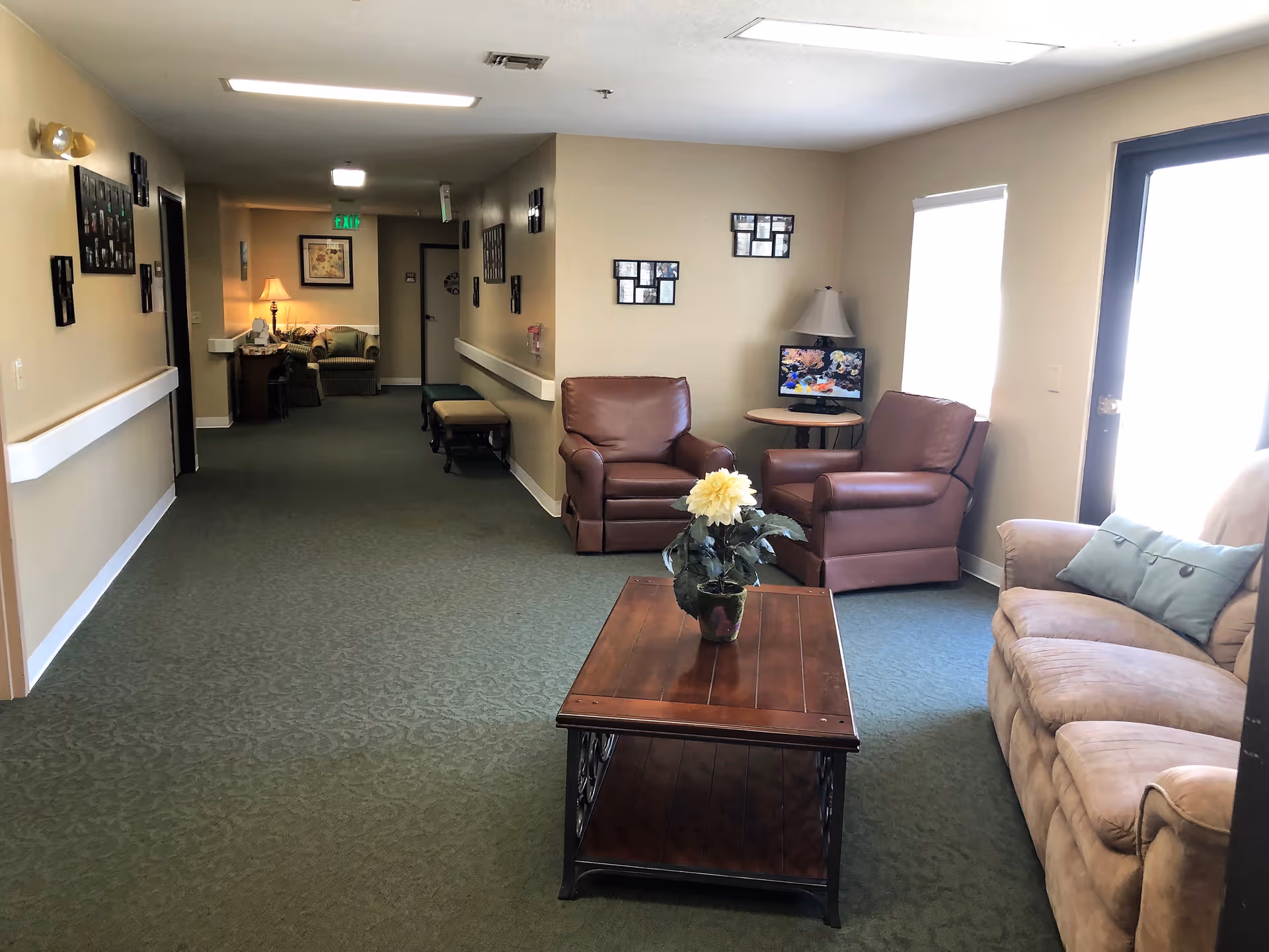 A cozy senior living facility common area with a green carpeted floor, beige walls, and ceiling lights. The room features a beige sofa with a blue pillow, two brown leather armchairs, a wooden coffee table with a potted plant, and a small round table with a lamp and a small TV. The hallway extends into the background with additional seating and framed pictures on the walls.