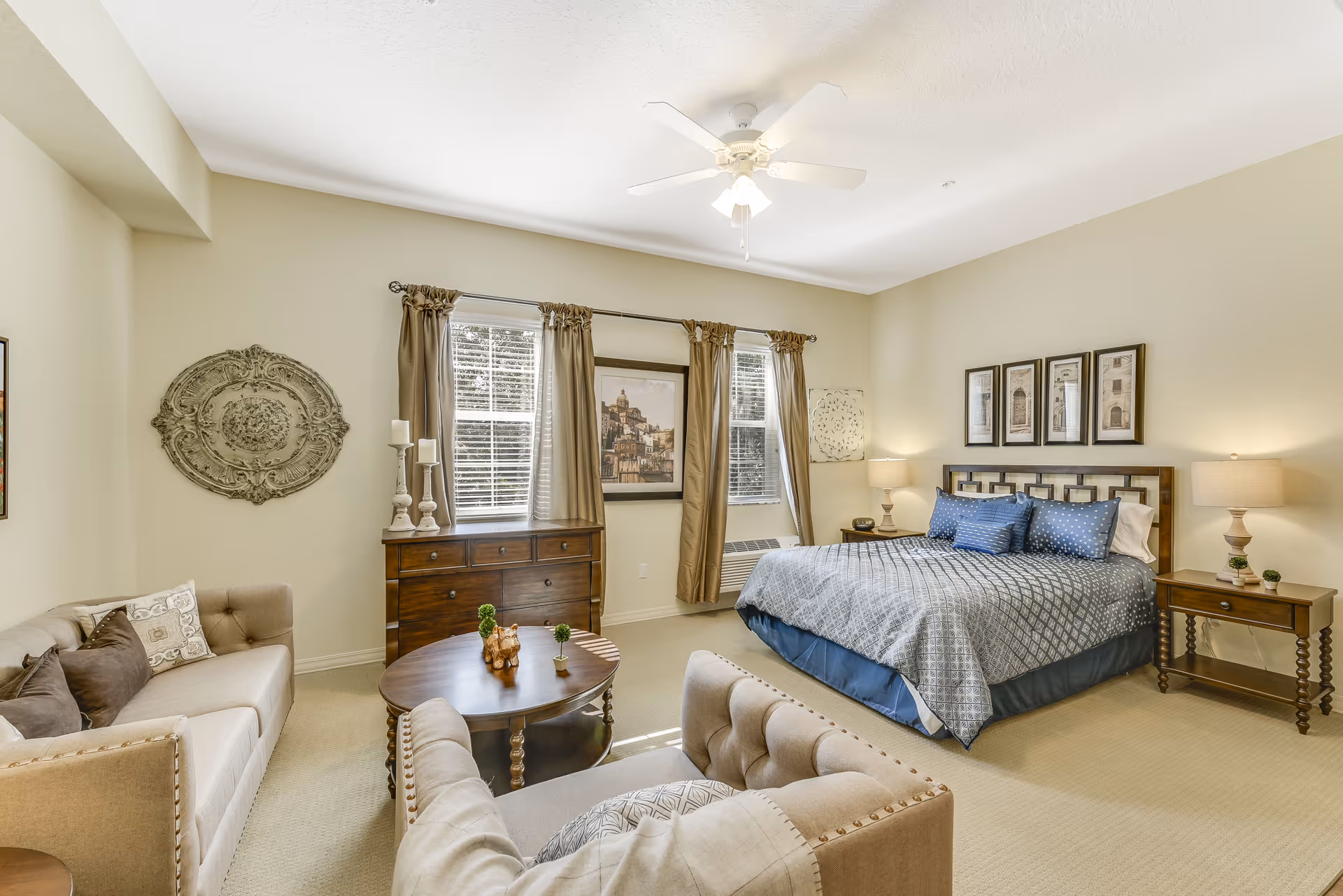A spacious and well-lit bedroom featuring a large bed with blue and gray bedding, two wooden nightstands with lamps, a wooden dresser under two windows with brown curtains, a round wooden coffee table with small plants, and two beige upholstered chairs and a sofa arranged around the table. The walls are decorated with framed artwork and a decorative wall piece.