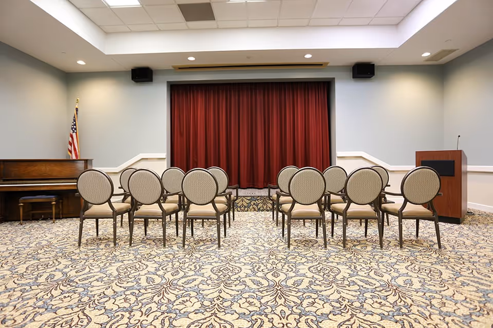 A small auditorium or meeting room with a patterned carpet, several rows of beige cushioned chairs facing a stage with a closed red curtain. To the left of the stage is an upright piano with a stool, and an American flag stands beside it. To the right of the stage is a wooden podium with a microphone.