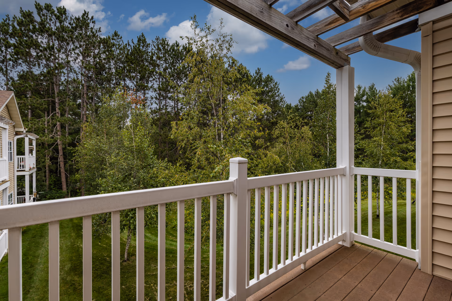 View from a balcony with white railing overlooking a green grassy area and trees under a partly cloudy blue sky. The balcony has a wooden floor and a wooden pergola overhead.