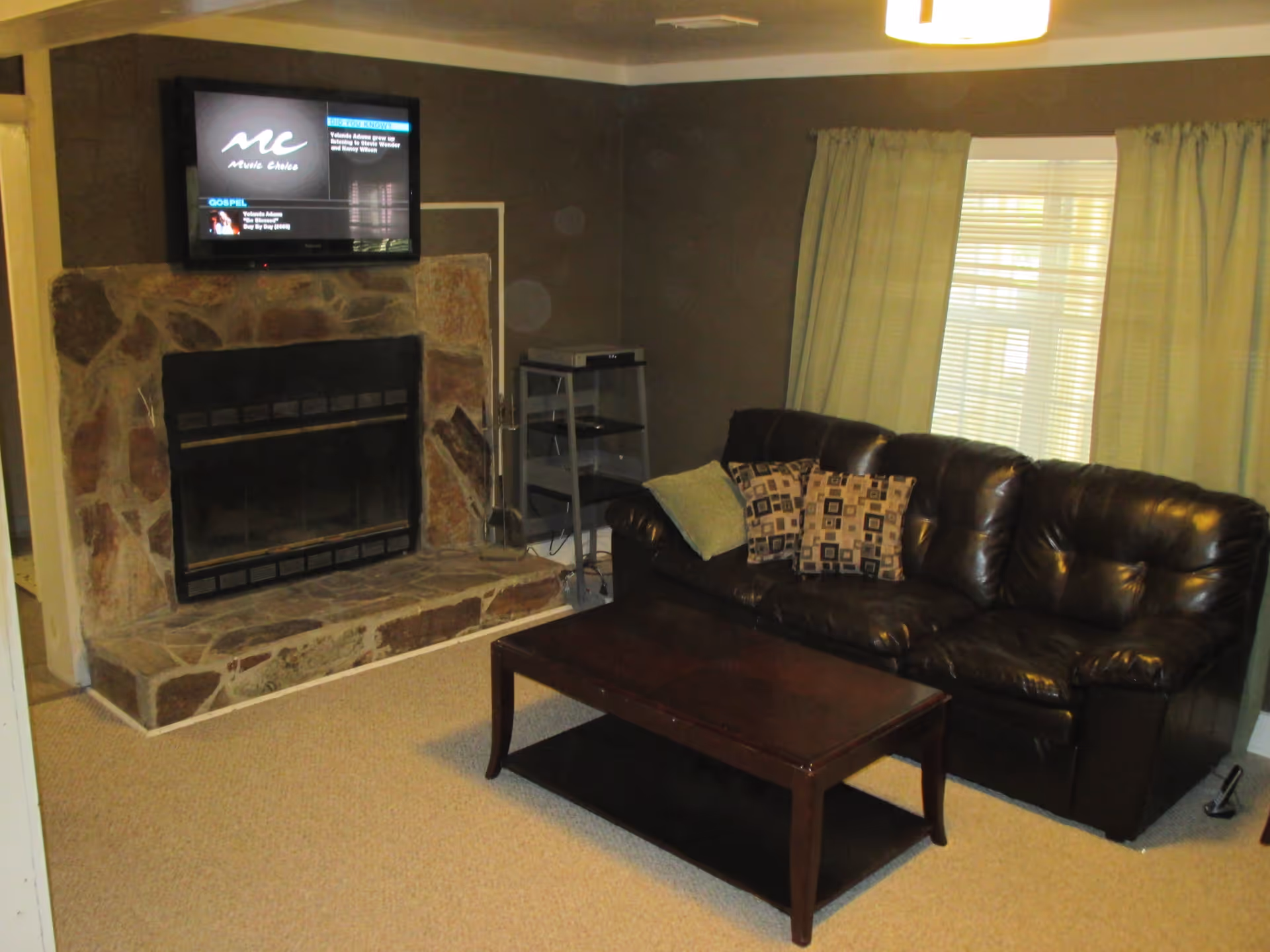 A cozy living room with a dark brown leather couch adorned with three patterned cushions, a wooden coffee table in front, a stone fireplace with a mounted flat-screen TV above it, and a window with light-colored curtains letting in natural light.