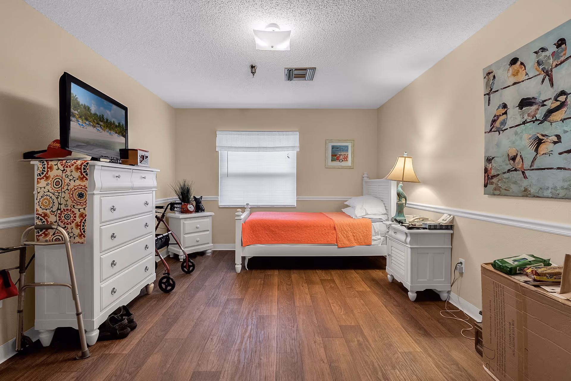A senior living bedroom with a single white bed covered with an orange blanket, a white dresser with a TV on top, a nightstand with a lamp, a walker, and a cardboard box on the floor. The walls are beige with white trim, and there is a window with blinds in the center. A painting of birds is hung on the right wall.