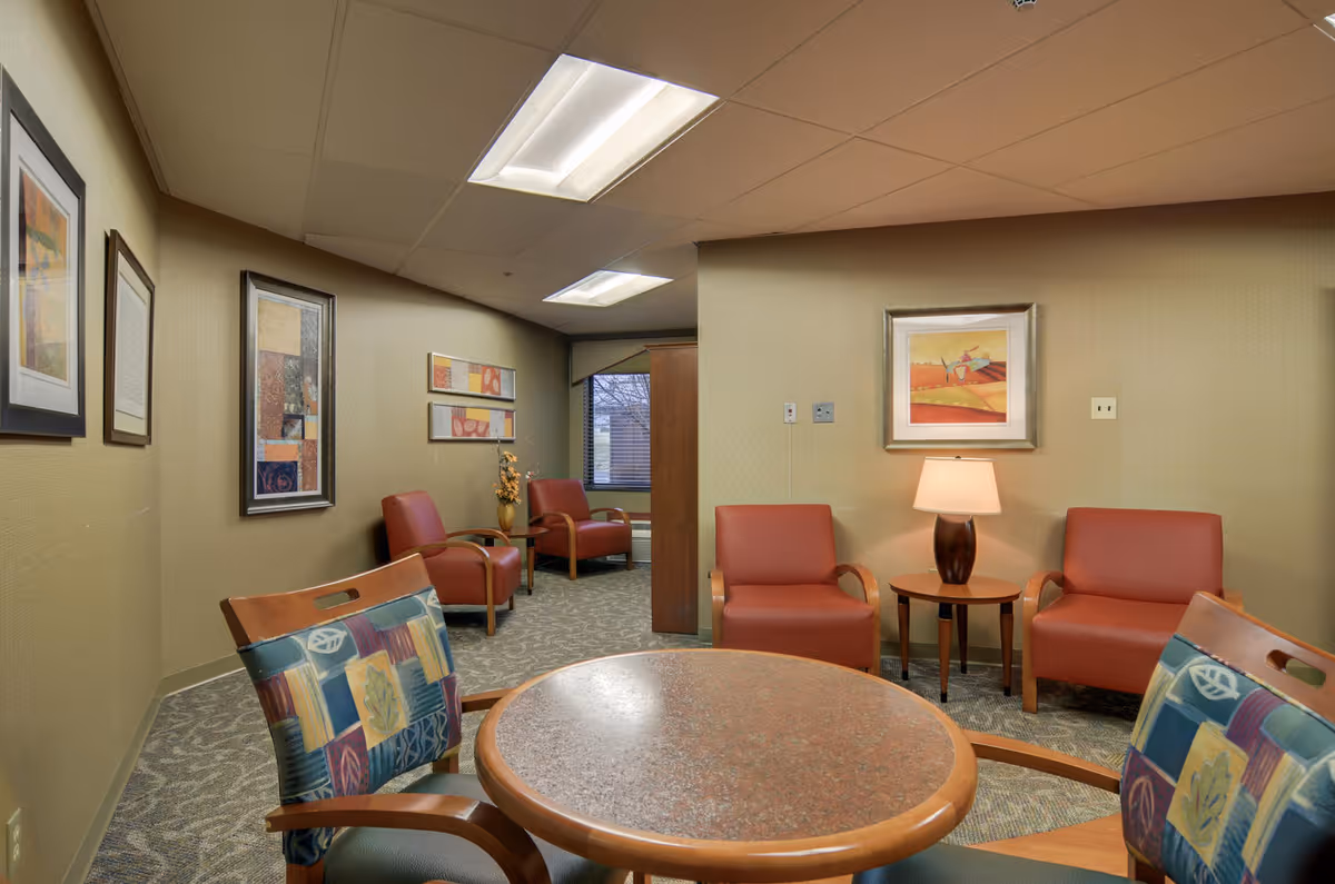 A cozy seating area in a senior living facility with four red armchairs arranged around a small wooden table with a lamp. There is a round table with two colorful cushioned chairs in the foreground. The walls are decorated with framed artwork, and a window is visible in the background.