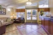 A cozy living room area in Ridgeway Post Acute featuring a patterned sofa, an armchair, a side table with a lamp, a floral painting on the wall, and large glass doors with plaid valances letting in natural light. There is a wooden reception desk with a flower arrangement on the right side.