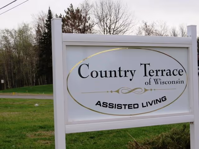 A white sign with gold and black lettering that reads 'Country Terrace of Wisconsin ASSISTED LIVING' standing on a grassy area with trees and a road in the background.